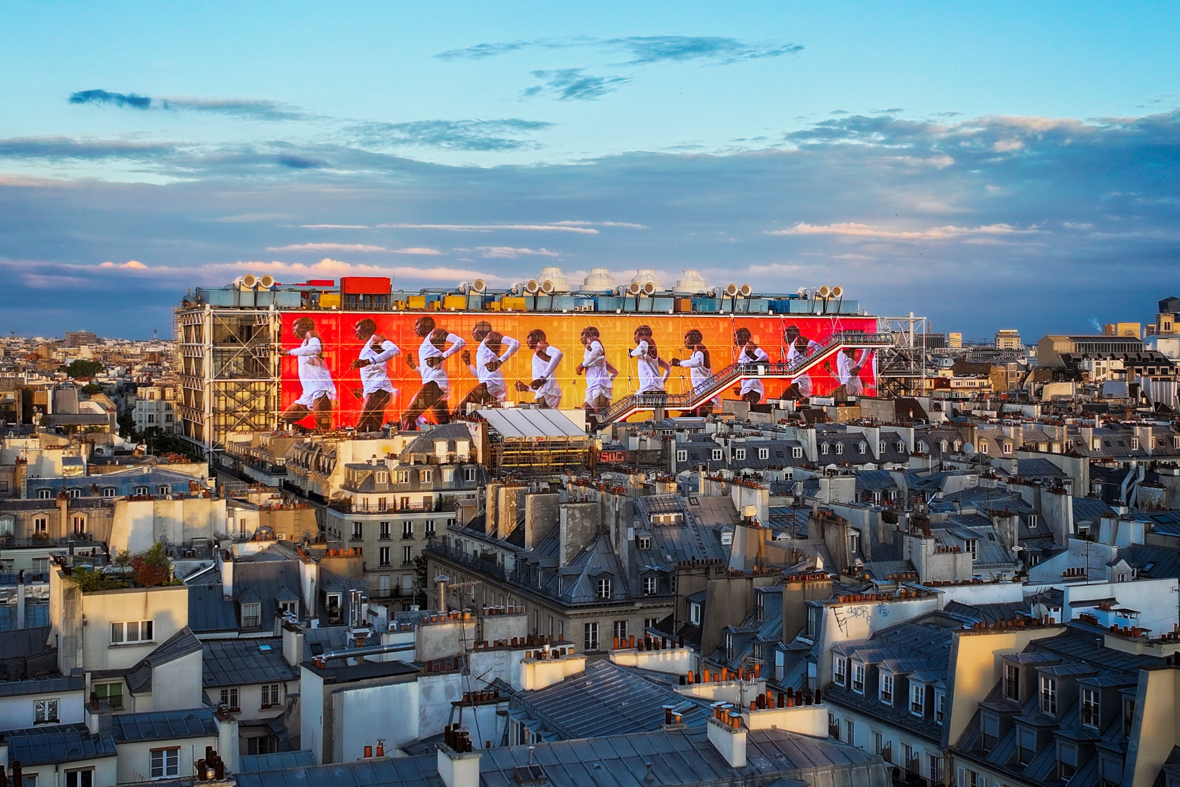 The transformed Nike Au Centre Pompidou’s iconic façade features a canvas that showcases groundbreaking stories of sport. Screen features repeating image of Eliud Kipchoge on bright red and yellow colors, which pops out against the cityscape and a cloudy blue Paris sky.