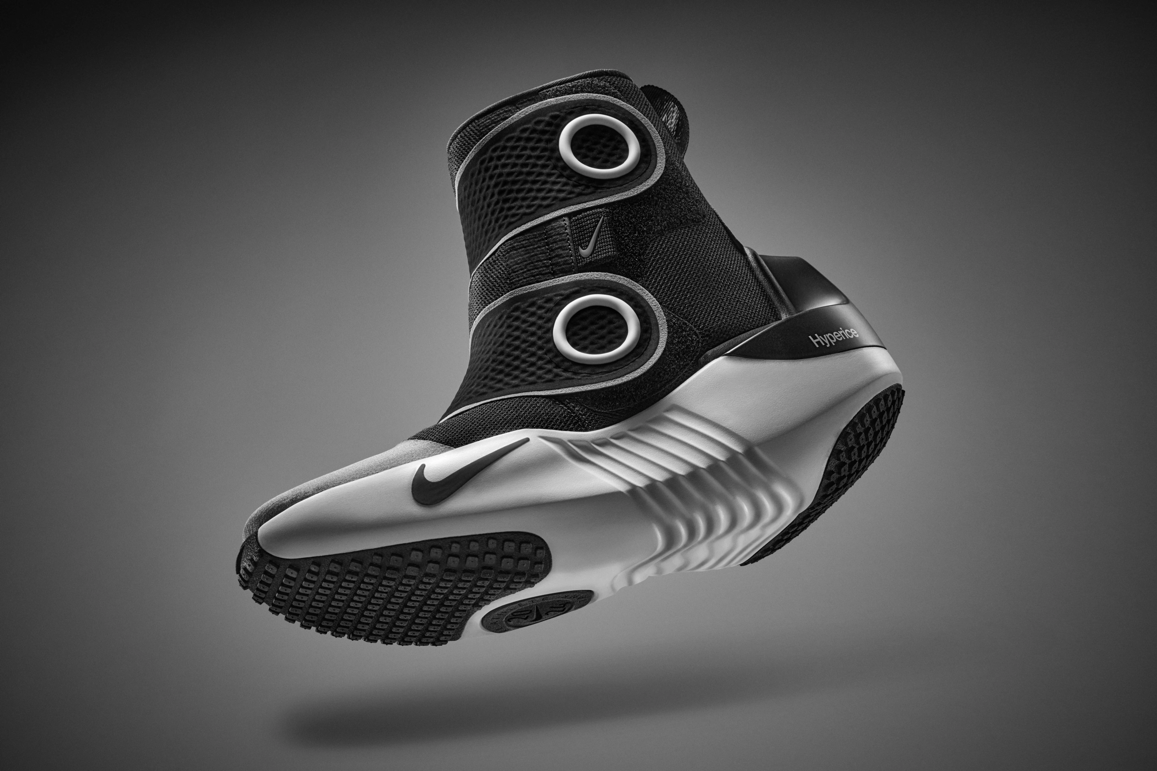 Front angled view of the Nike x Hyperice boot, which is a high-top boot with a black upper, white midsole, and black and white sole. The boot's closure system has large black velcro tabs with white circles. The boot floats on a grey background with a slight shadow beneath.