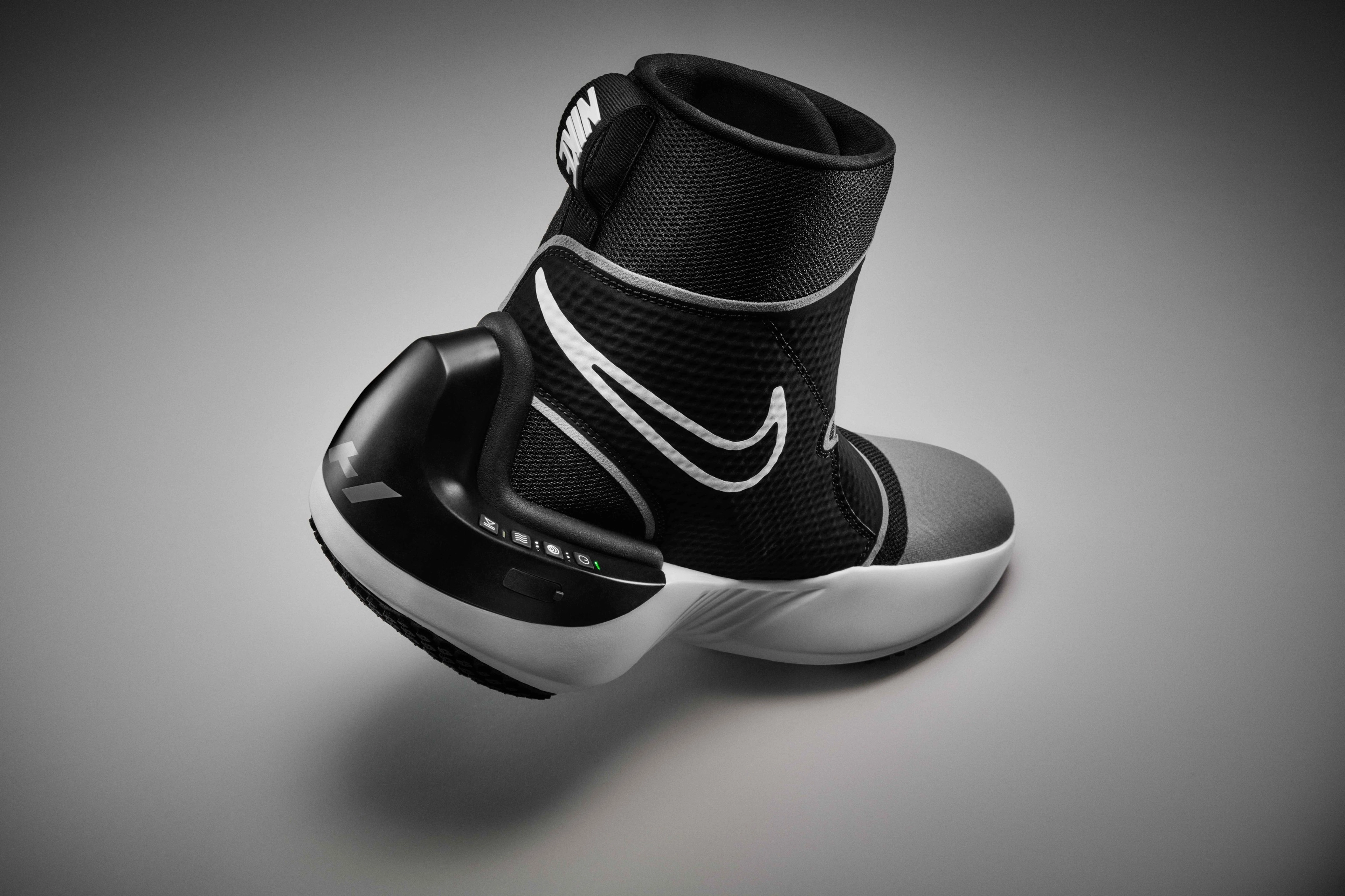 Back view of the Nike x Hyperice boot, which is a high-top boot with a black upper, white midsole, and black and white sole. We see the control unit for the boot, with a number of control buttons. The boot's toe sits on a grey floor, while the heel floats above, casting a shadow underneath.