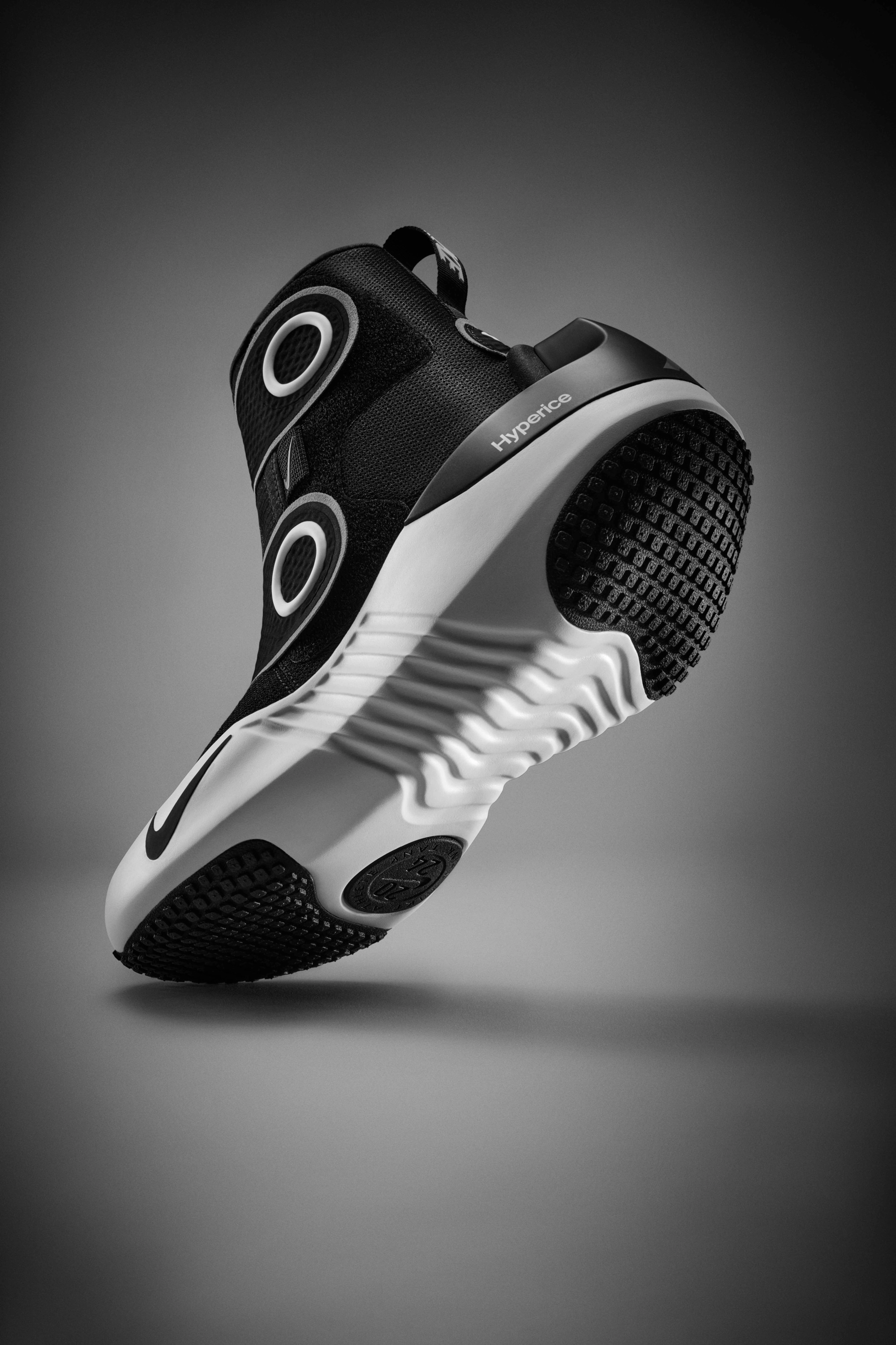Underside view of the Nike x Hyperice boot, which is a high-top boot with a black upper, white midsole, and black and white sole.  We see black waffle pattern pods at the toe and heel of the sole. The boot's toe sits on a grey floor, while the heel floats above, casting a shadow underneath.