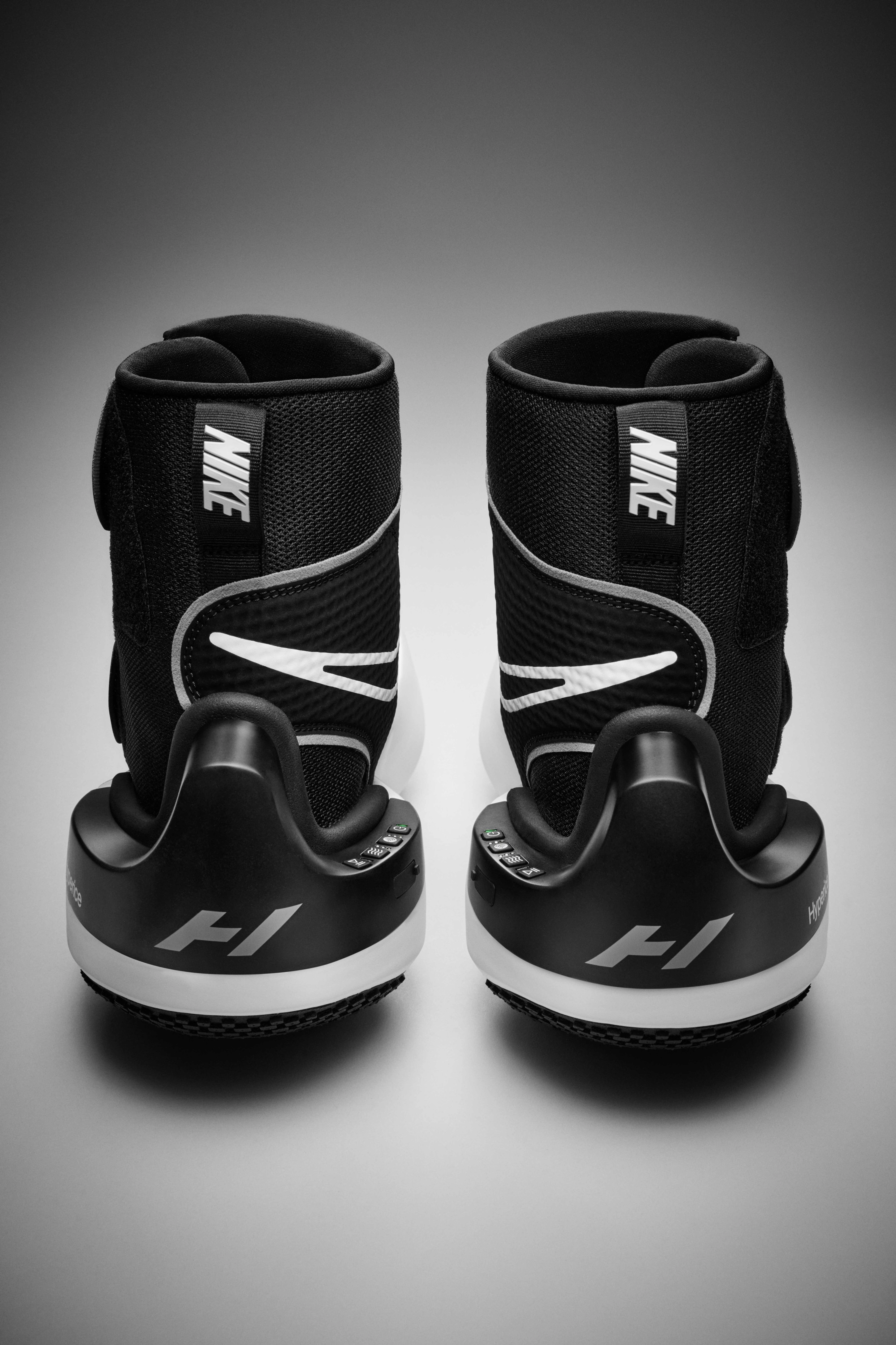 Back view of a pair of Nike x Hyperice boots, detailing the control units on each shoe.  Black boots sit on a grey surface.
