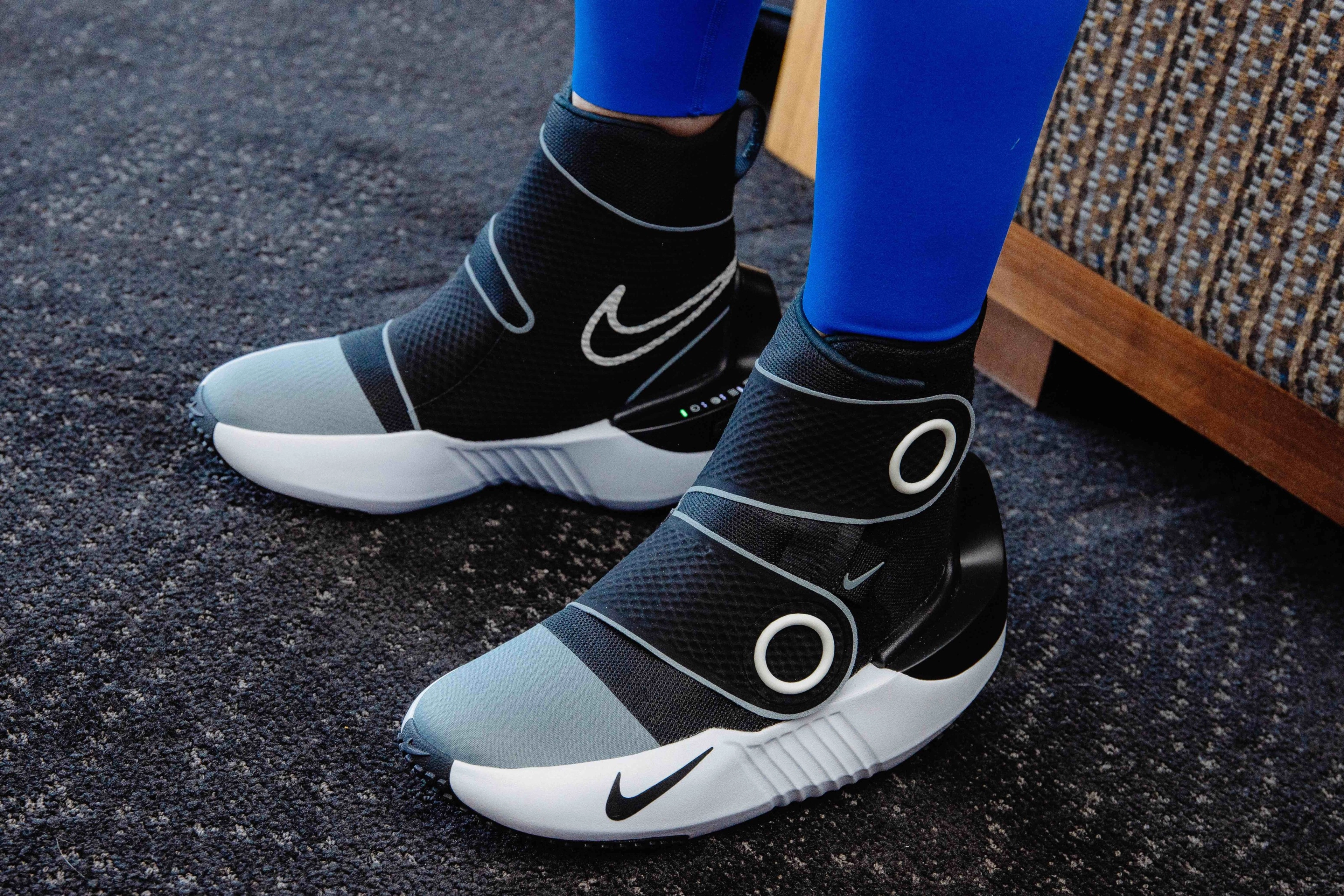 Left close up view of an athlete's feet in the Nike x Hyperice boots. Athlete wears bright blue tights with black boots, while standing on dark grey patterned carpet.