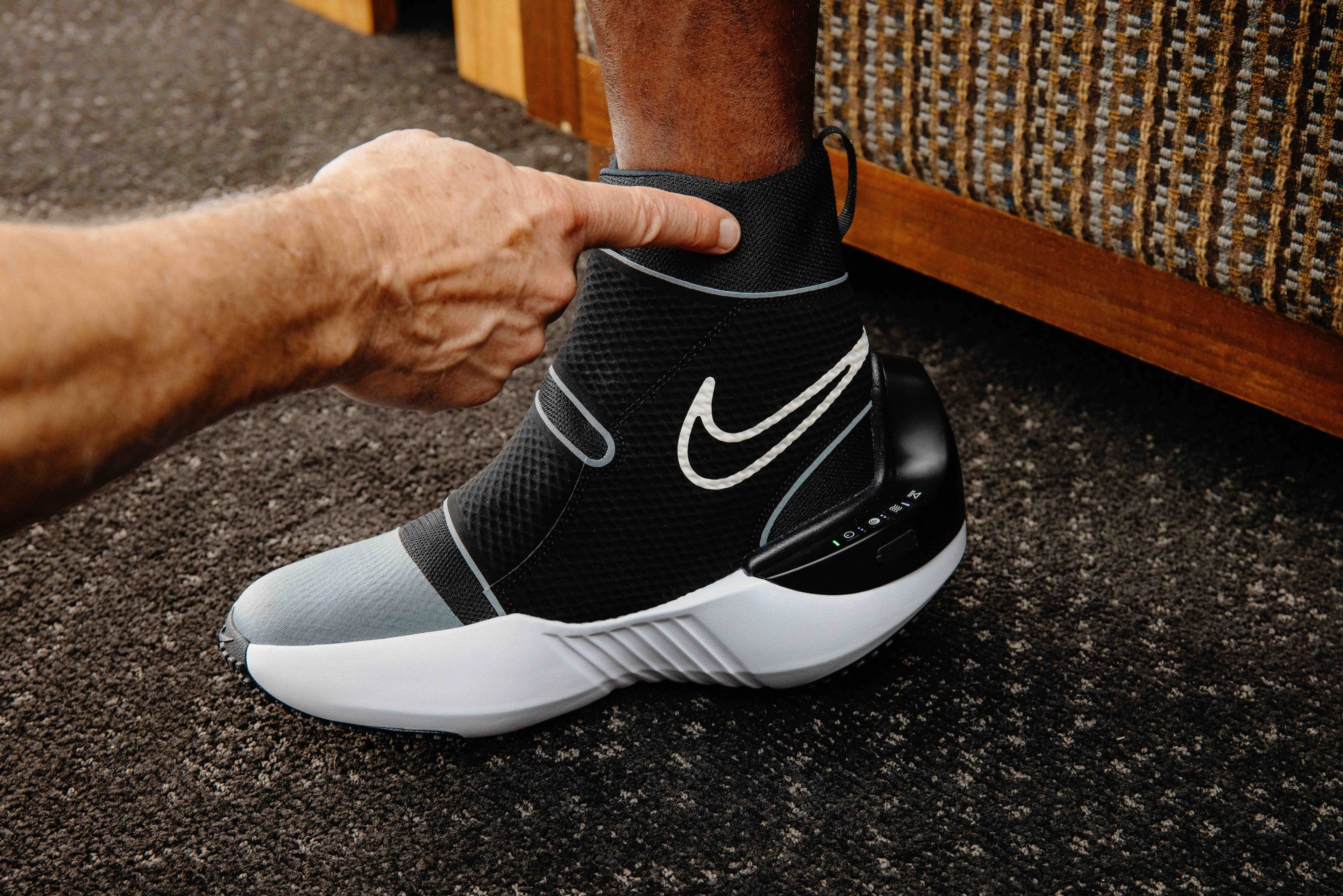 Left view of Nike x Hyperice boot worn by an athlete. A hand points to the compression band at the top of the boot, as athlete stands on dark grey patterned carpet.
