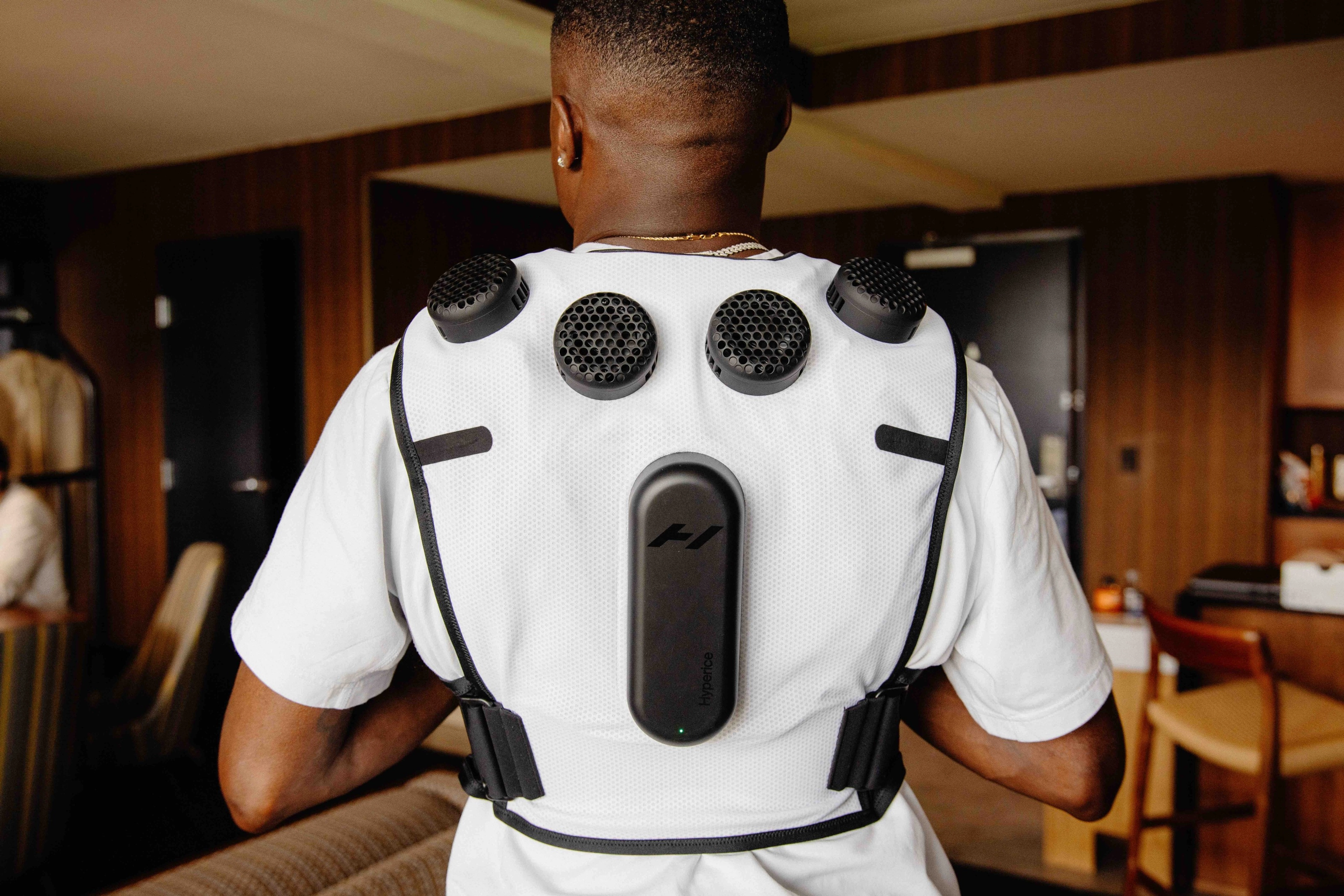 Back view of an athlete wearing the white Nike x Hyperice vest over a white tee shirt. The athlete's back is facing the camera, so we see the black thermal modules on the vest unit.