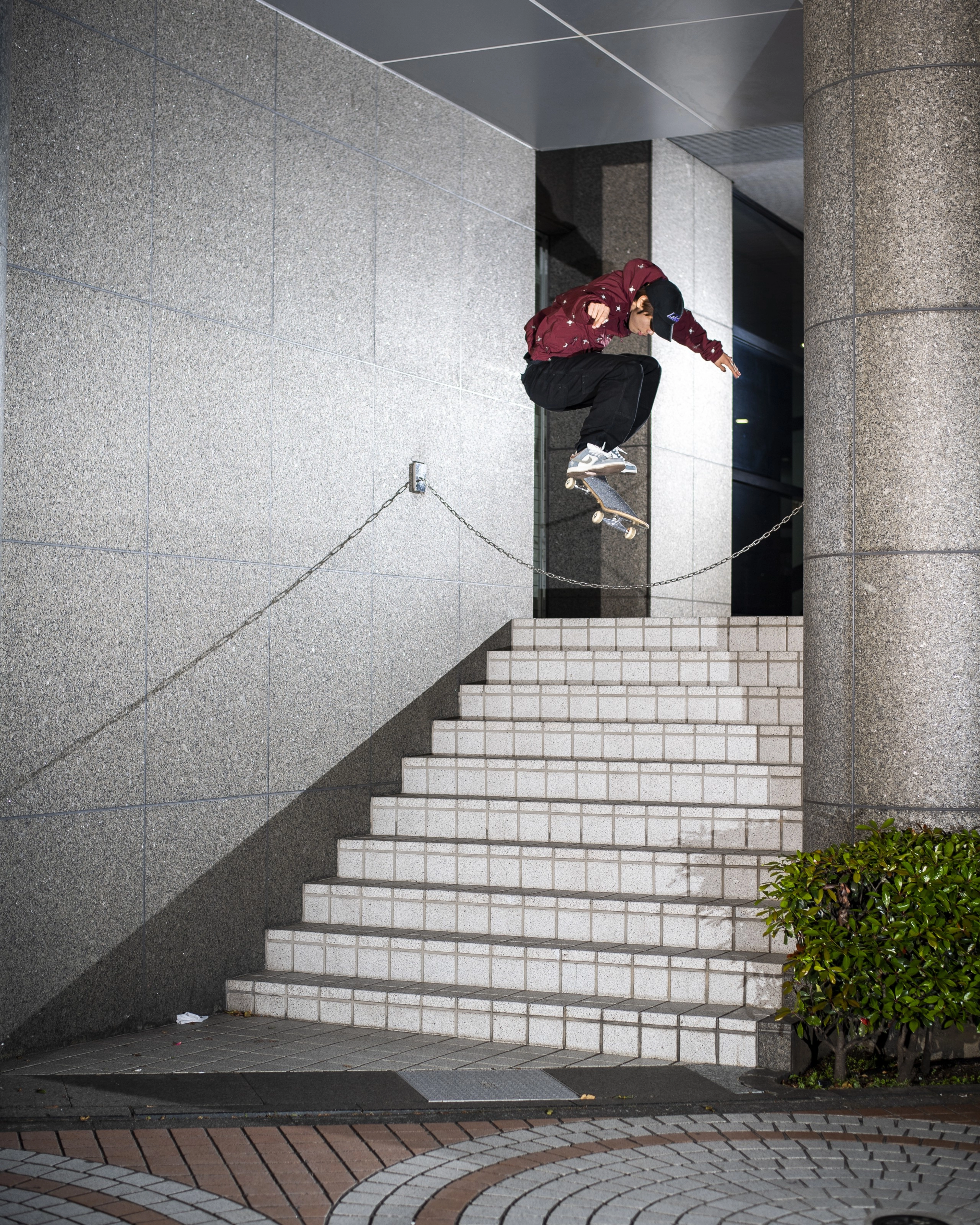Yuto Horigome jumps a chained off stairwell on his skateoard. He wears black pants, a maroon hoodie, and black Nike cap, with grey and white Nike SB Dunks. The stairs are white cement against a granite wall.