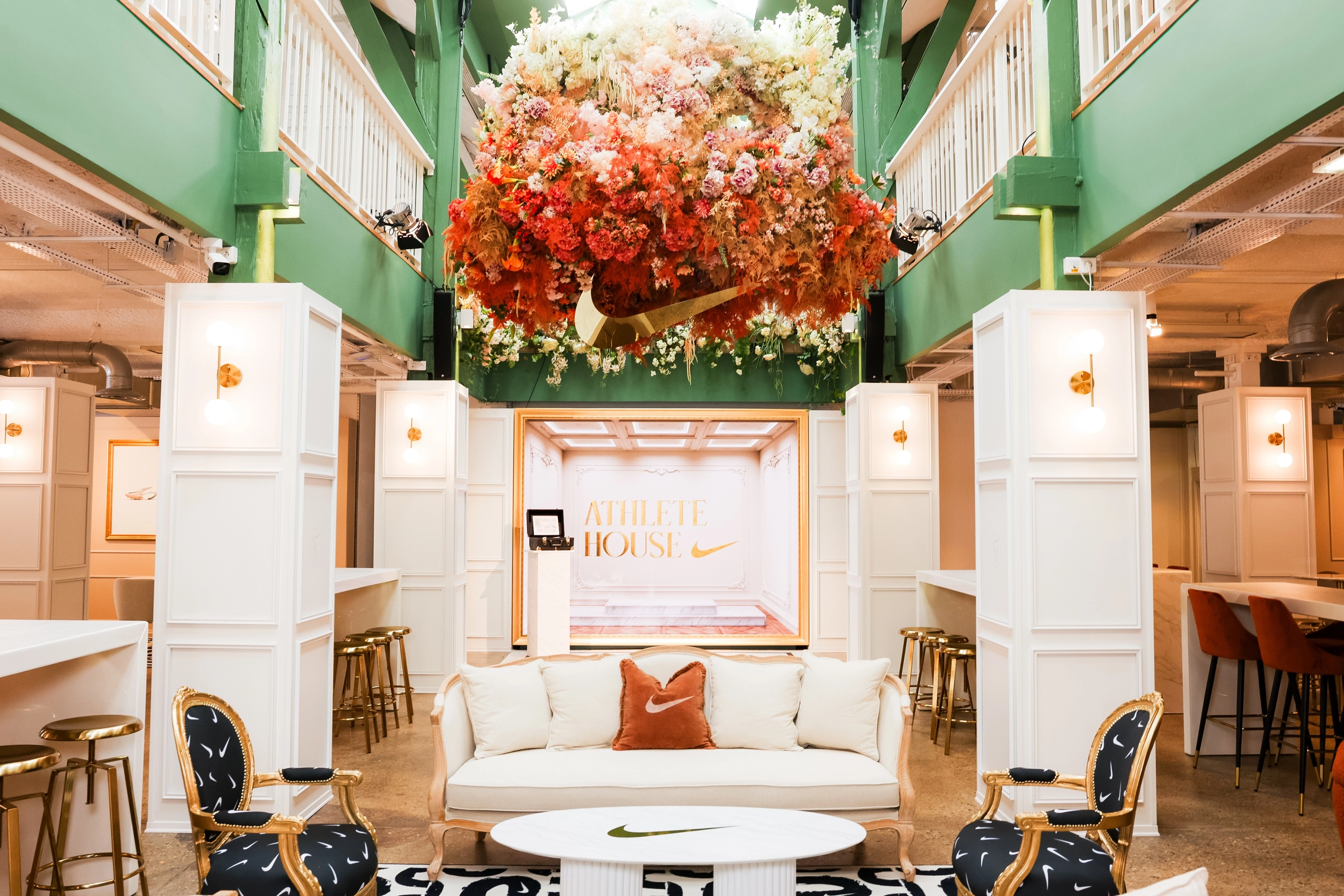A front view into the entryway of the Nike Athlete House in Paris, France. A dramatic flower display with a gold Swoosh hangs above a lounge space with green walls. 2 chairs are upholstered with black fabrics with white Swooshes. A white sofa holds a rust colored pillow with a white Swoosh. There is white cabinetry and high tables with barstools flanking the room.