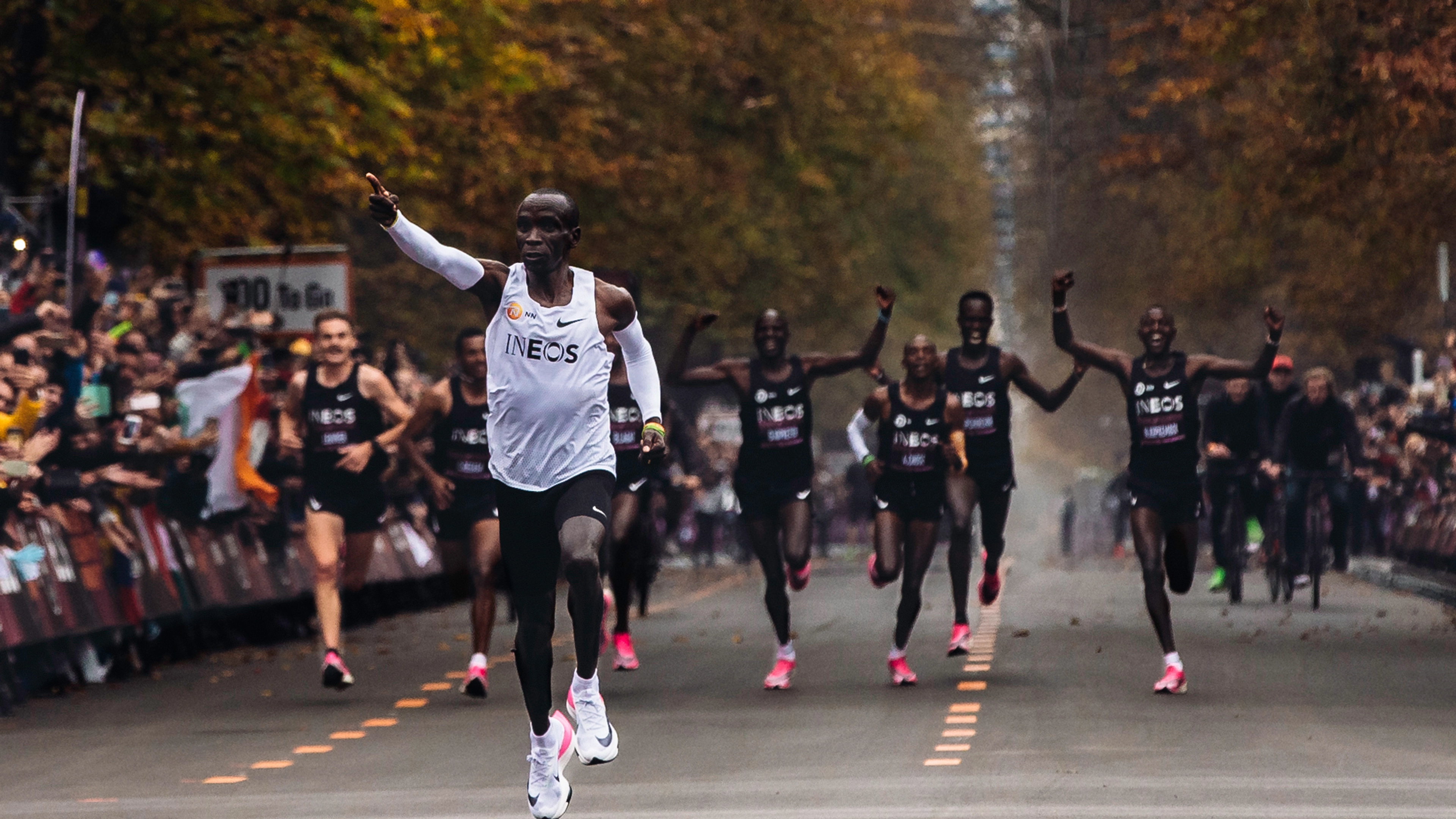 Eliud Kipchoge wearing Nike Air running sneakers and pointing forward as he leads a running marathon.