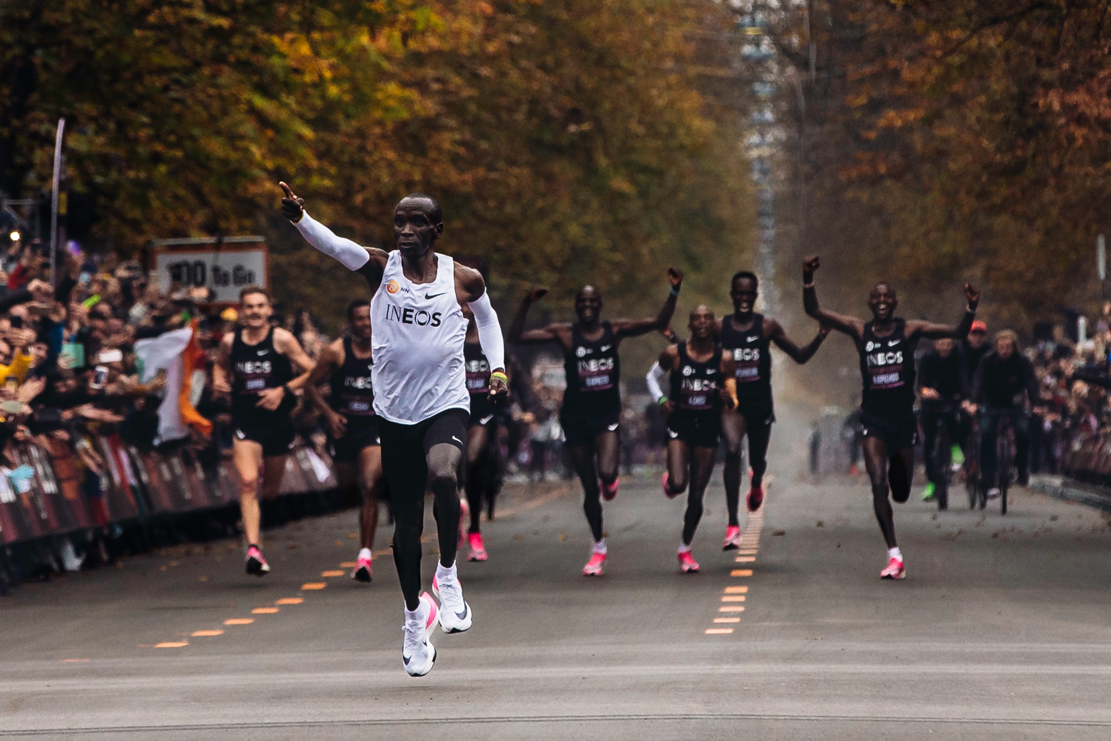 Eliud Kipchoge wearing Nike Air running sneakers and pointing forward as he leads a running marathon.