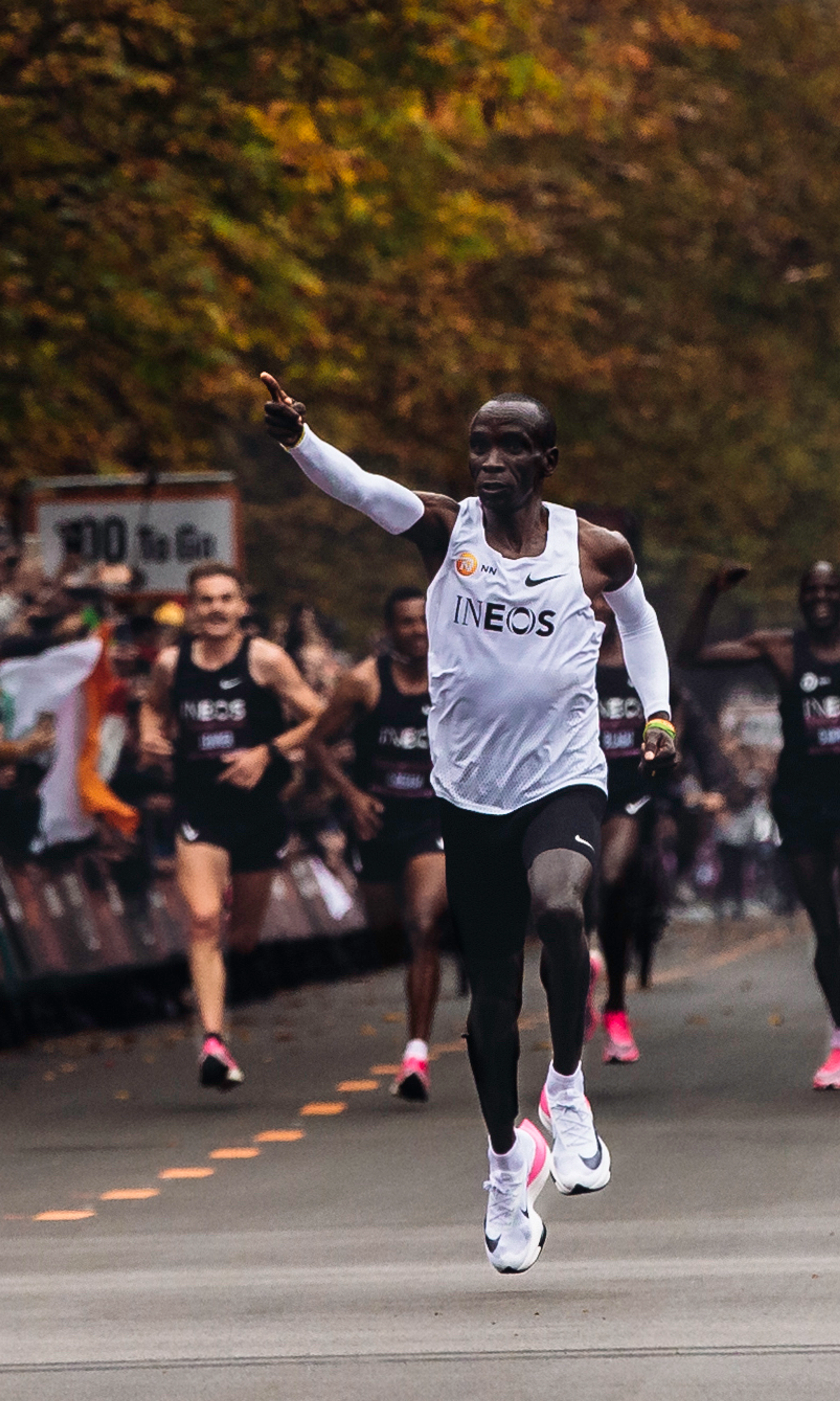 Eliud Kipchoge wearing Nike Air running sneakers and pointing forward as he leads a running marathon.