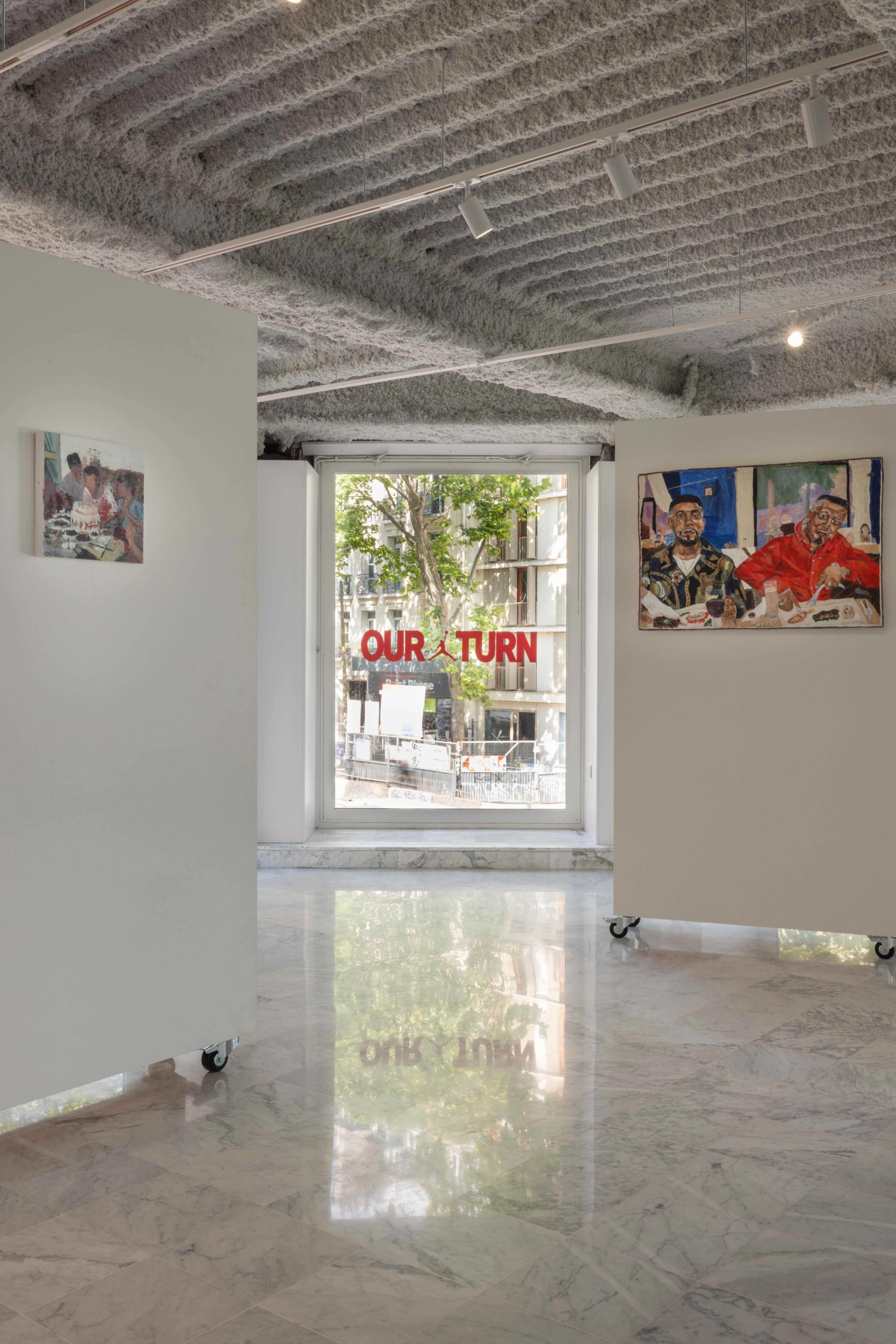 A photo of the District 23 gallery room featuring some artistic works on rolling displays and a window with "Our turn" printed on it behind them.