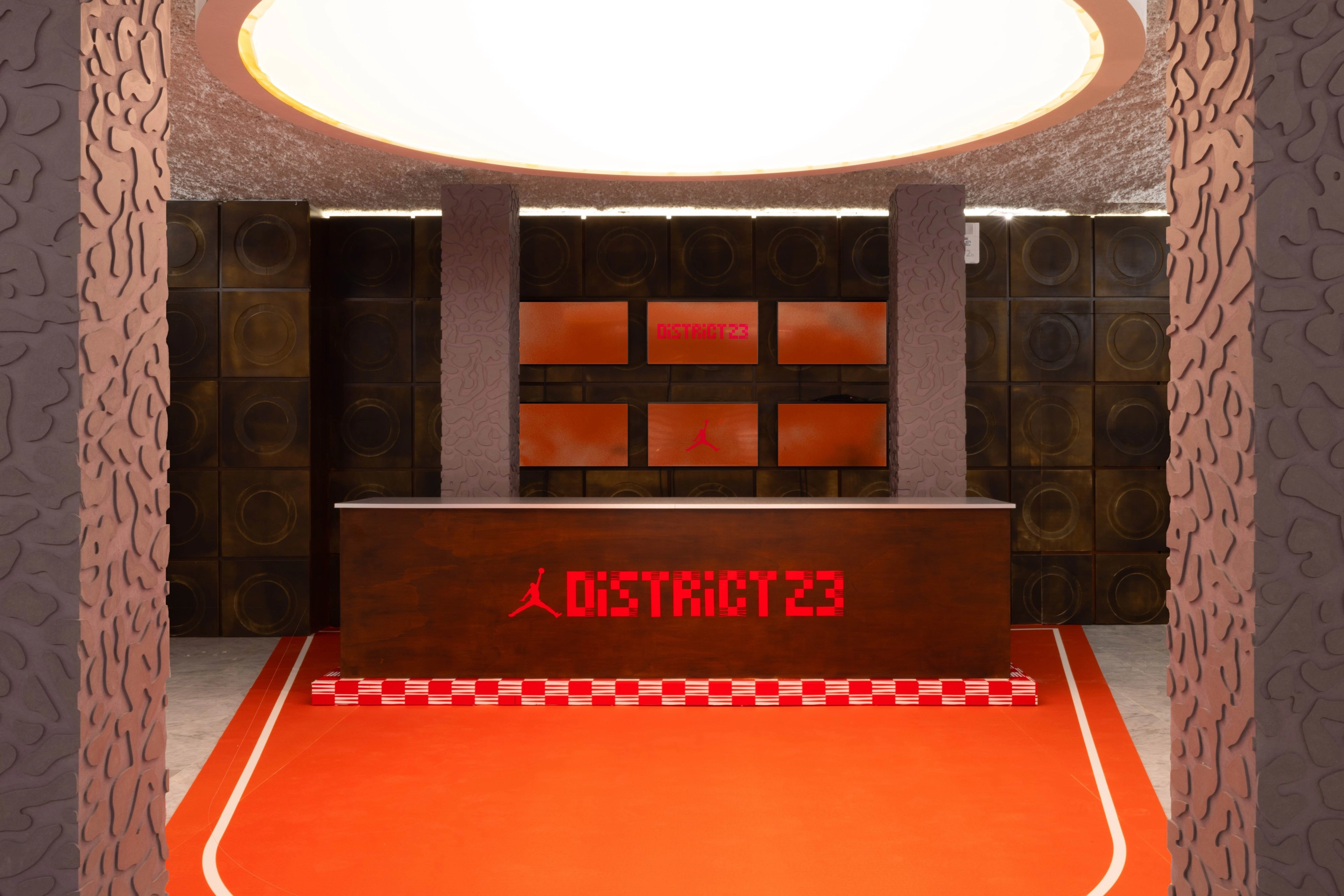 A photo of the District 23 main entrance area, featuring a front desk, orange flooring, a circular overhead light, and digital displays behind the desk.