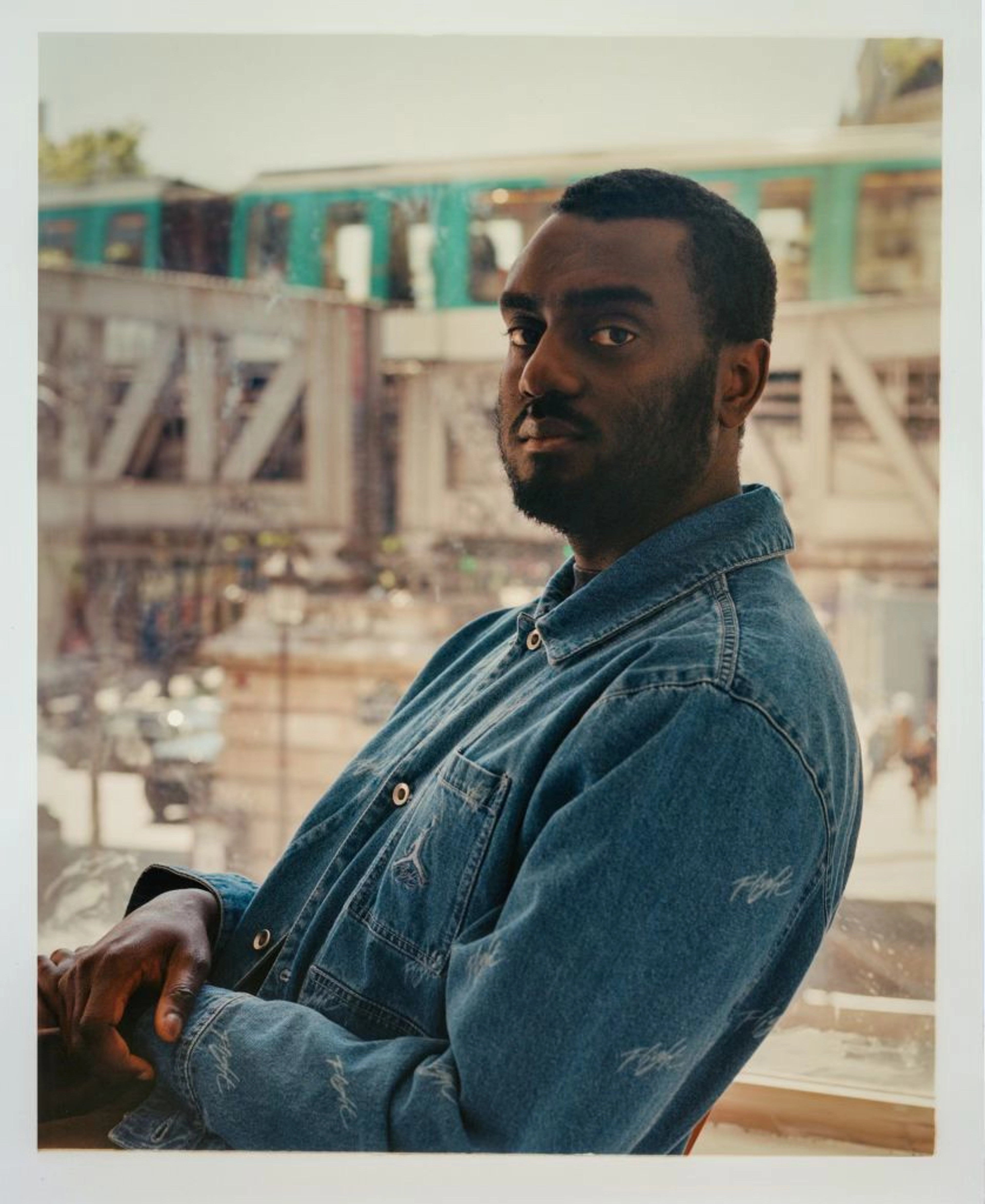 Youssouf Fofana sitting in front of a window with a passenger train passing by in the background.