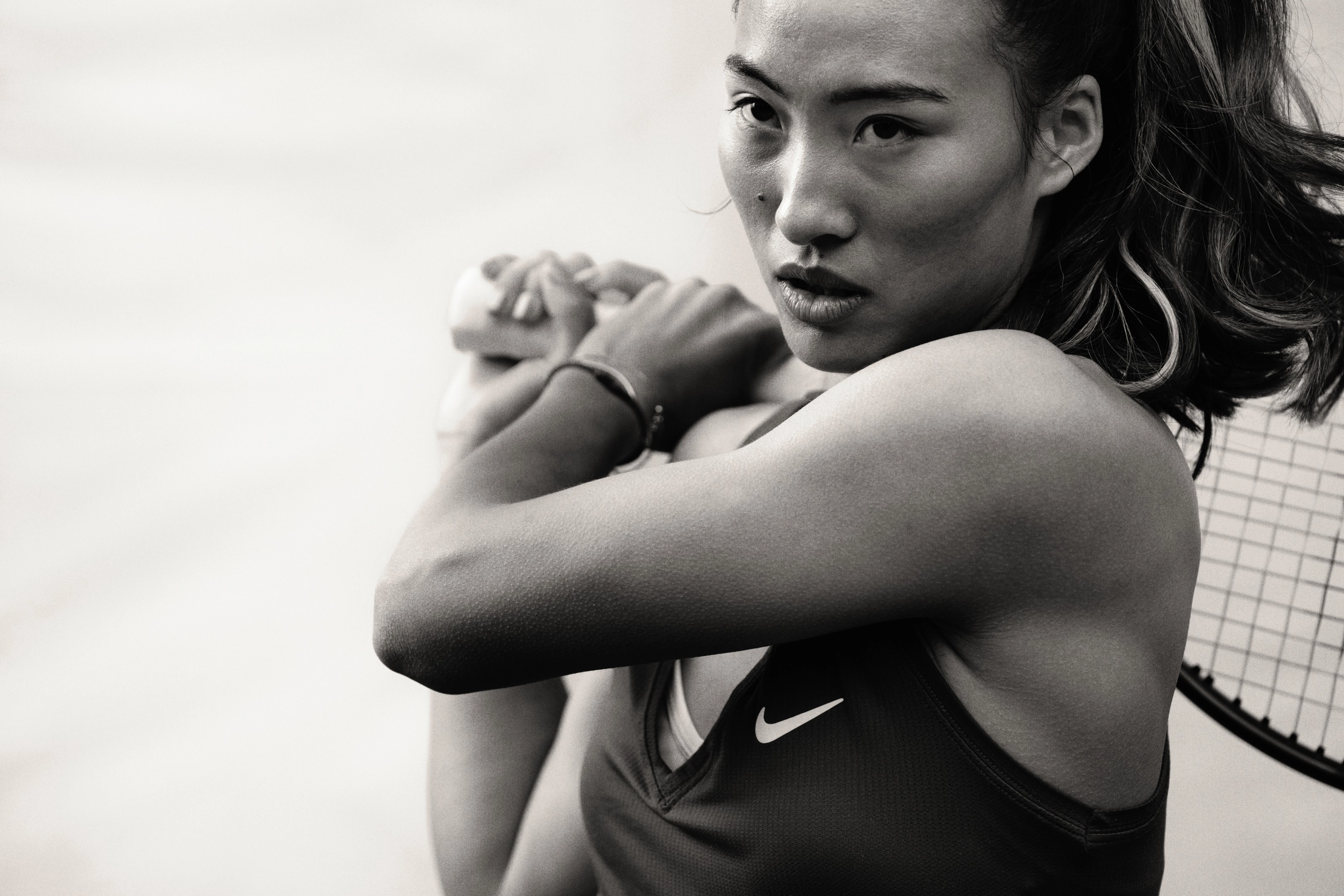 Black and white close up photo of Qinwen Zheng holding a tennis racquet over her shoulder with both hands. She wears a black NikeCourt Victory tank with a white Swoosh.