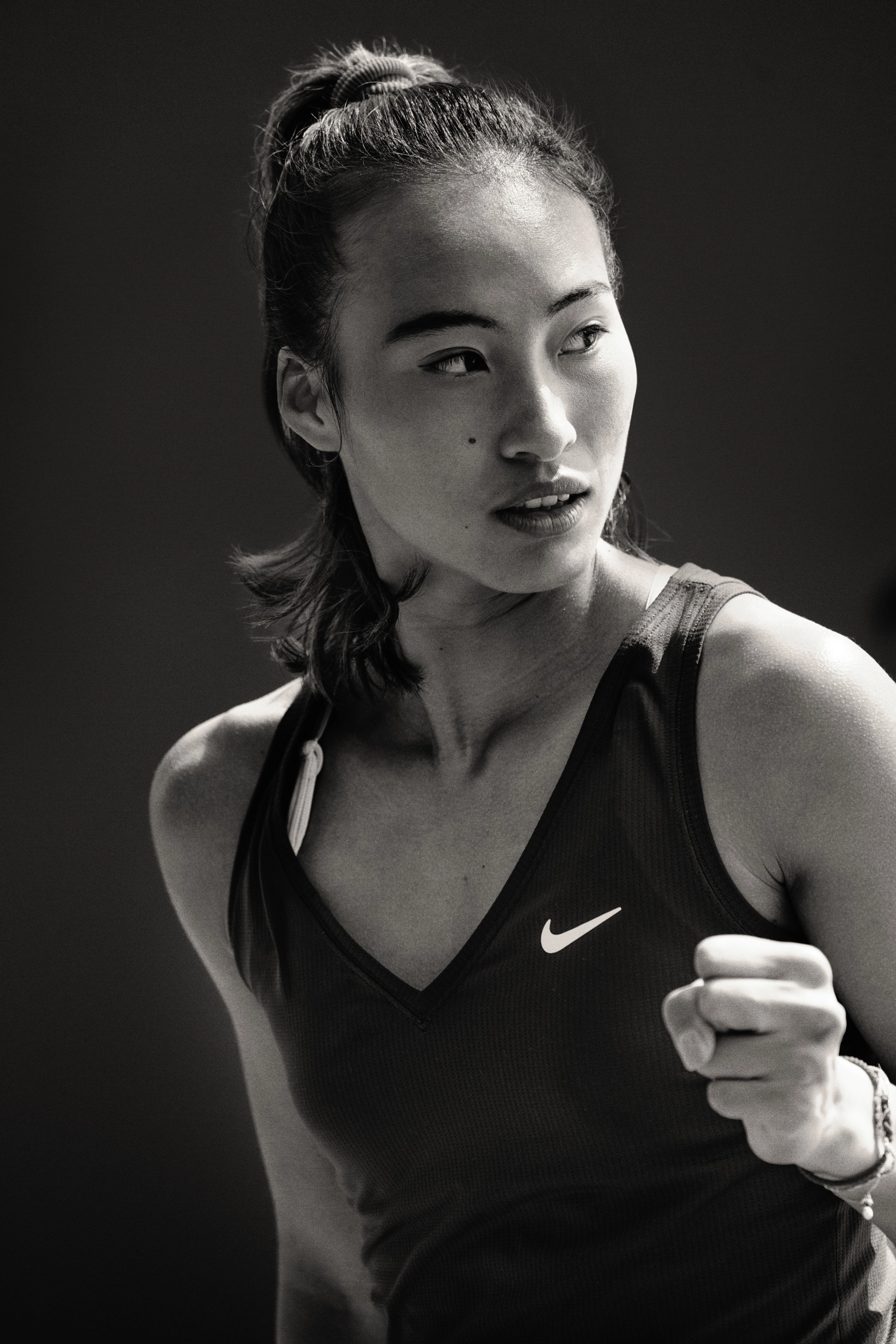 Black and white close up photo of Qinwen Zheng, wearing the NikeCourt Victory Tennis Tank in black. She looks across the tennis court and holds her hand in a fist.
