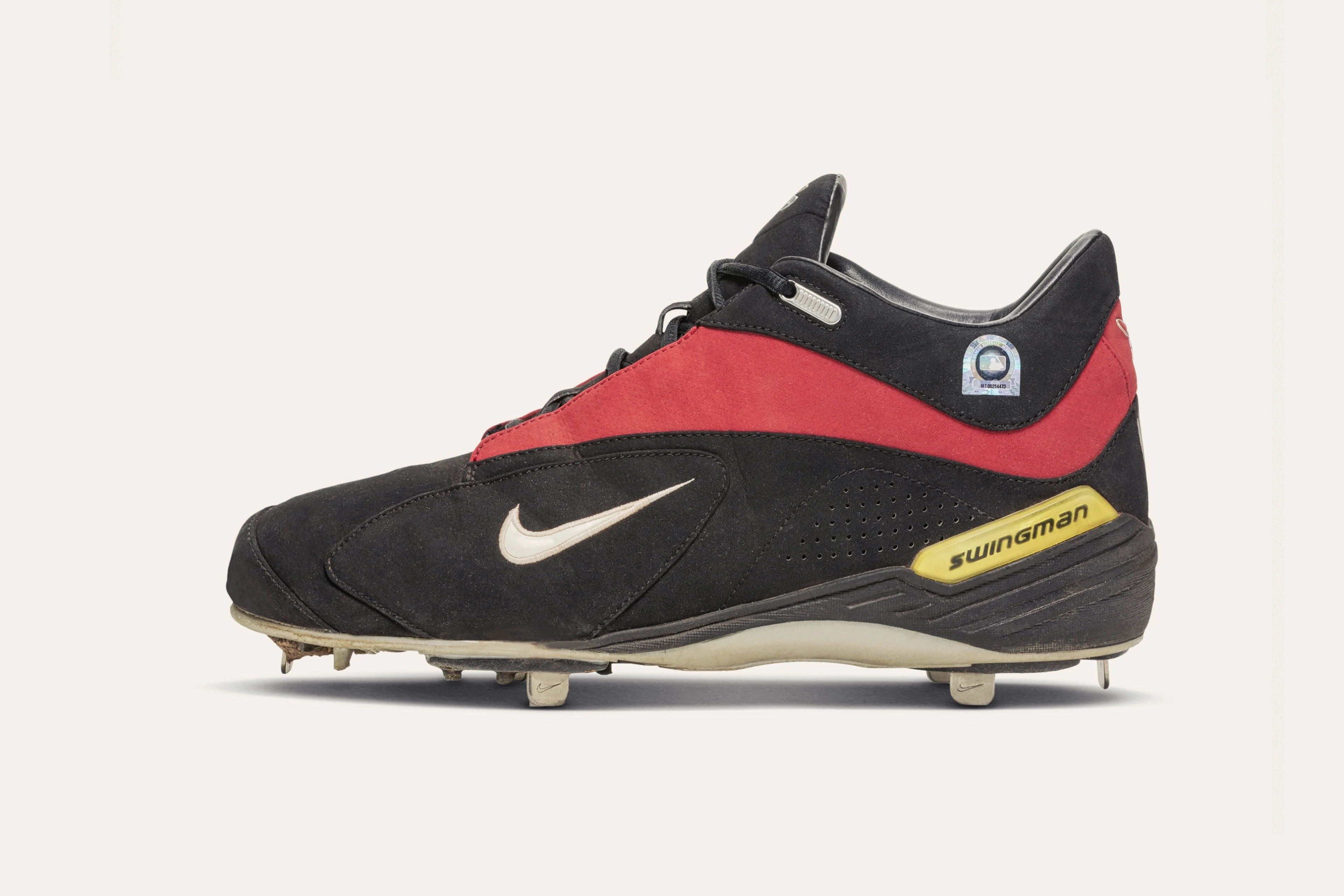 A dark grey sneaker with a small white Nike swoosh on the side, a red stripe around the ankle, a yellow tab on the heel that says "Swingman," and a white sole with cleats.