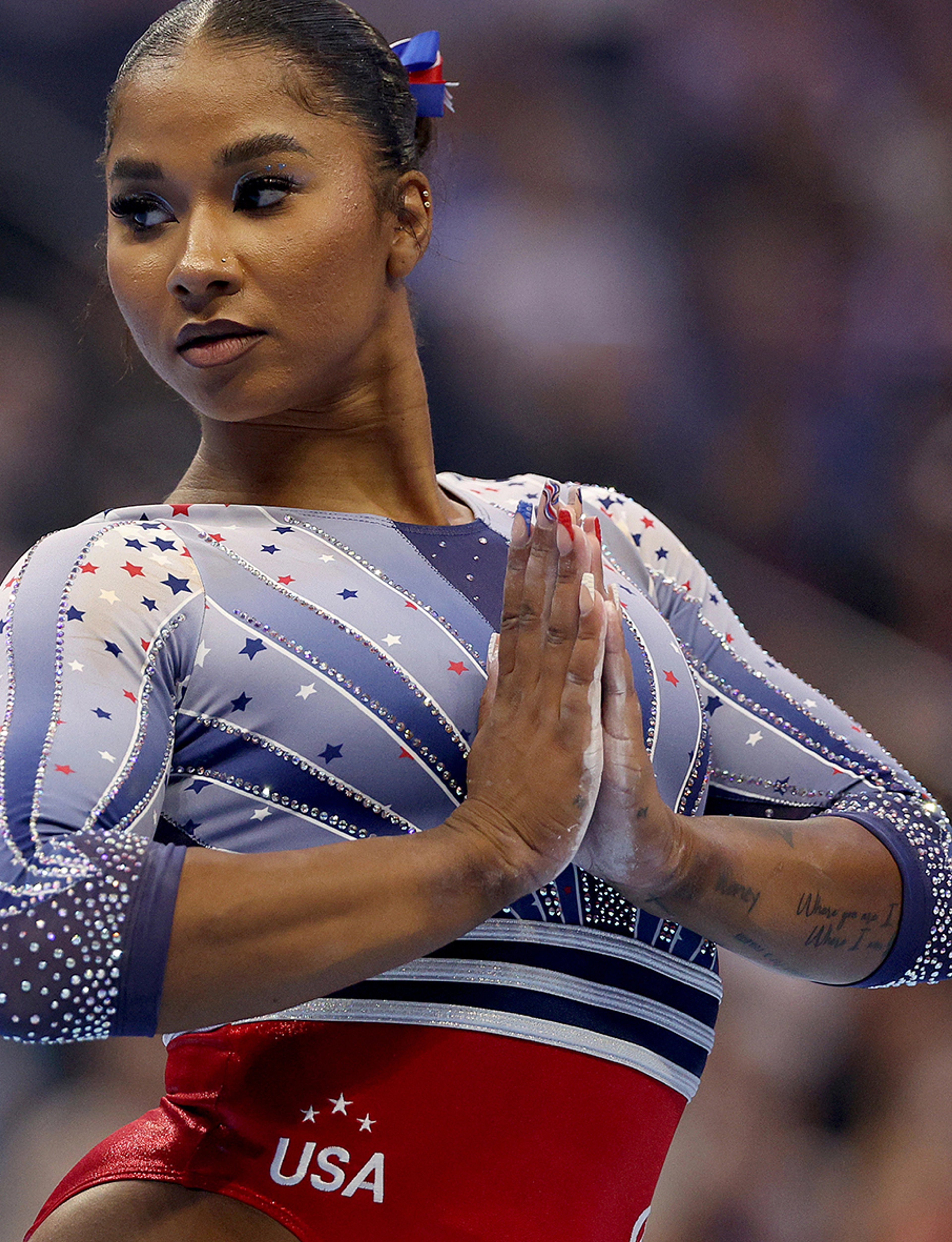 A close up of Jordan Chiles of Team USA Women’s Gymnastics taken while she is in competition. She looks to the left of camera and wears the Team USA one piece in red, white, and blue. She presses her chalk covered hands together in front of her chest.