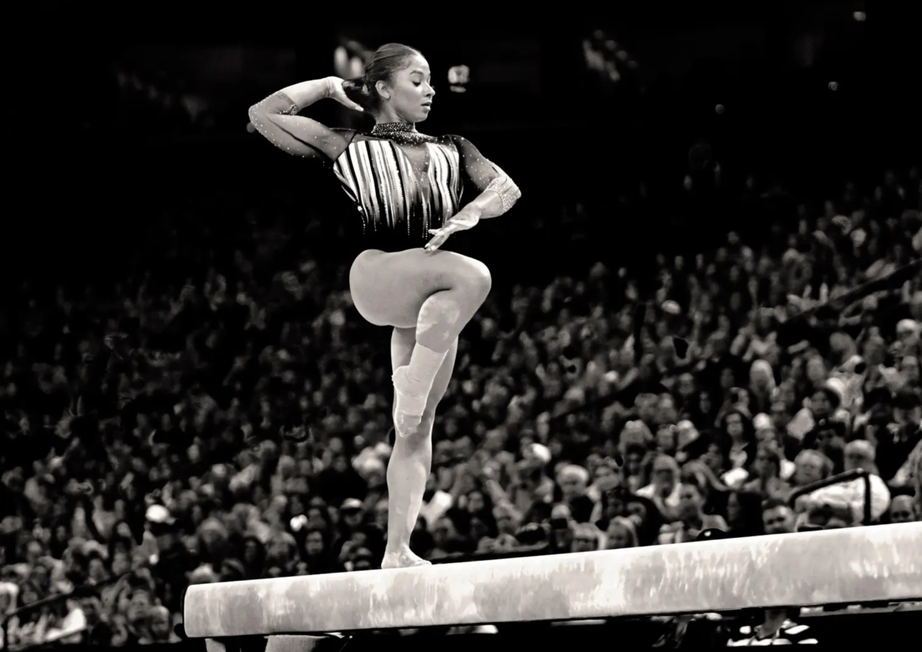 A black and white photograph of Jordan Chiles of Team USA Women’s Gymnastics taken while she's in competition on the balance beam. With a full crowd behind her, she lifts her knee and looks to her right.