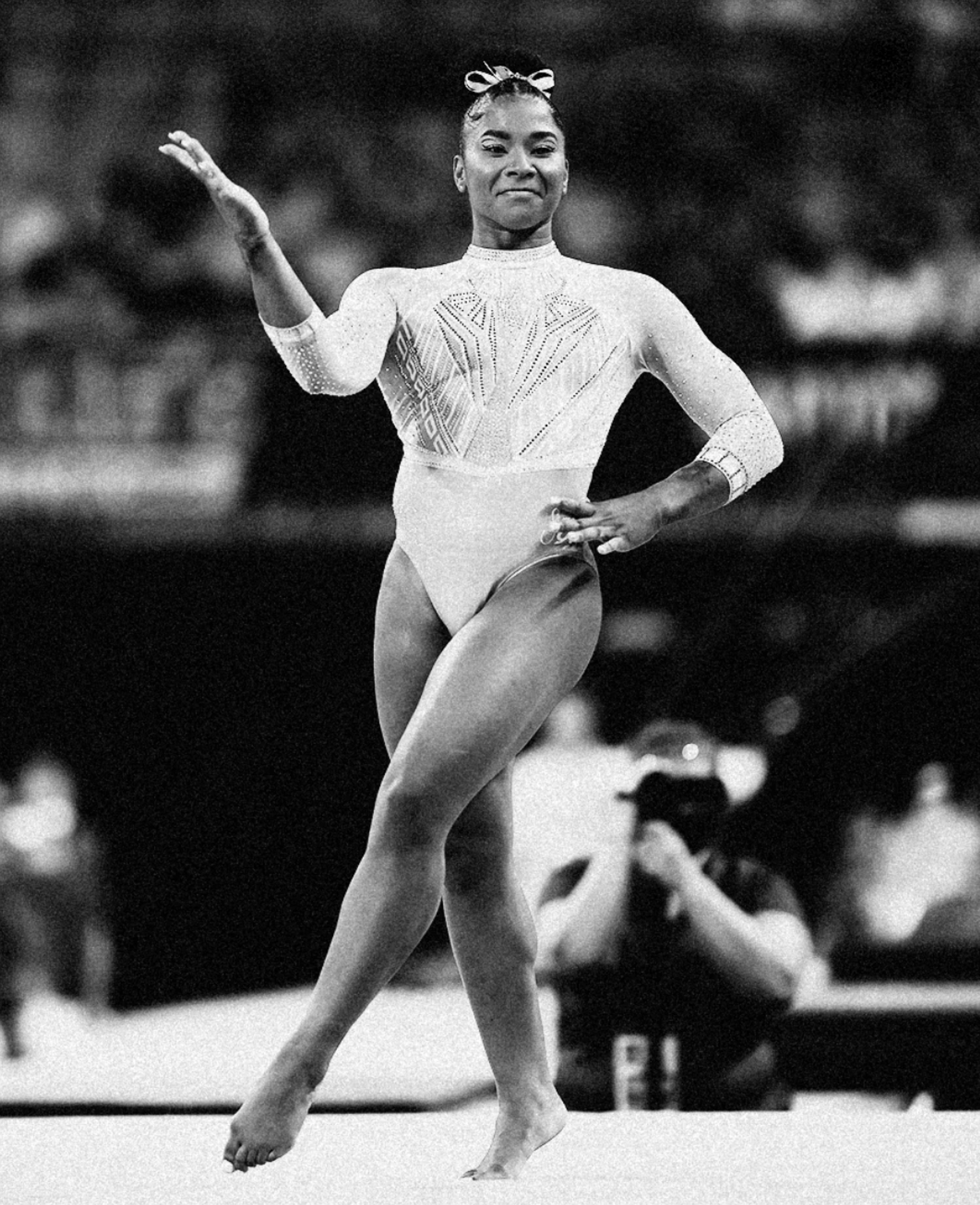 Black and white photo of Jordan Chiles of Team USA Women’s Gymnastics taken while in competition during the floor exercise. She crosses her leg as she moves on the mat, and raises her left hand above her shoulder. She smiles towards the camera.