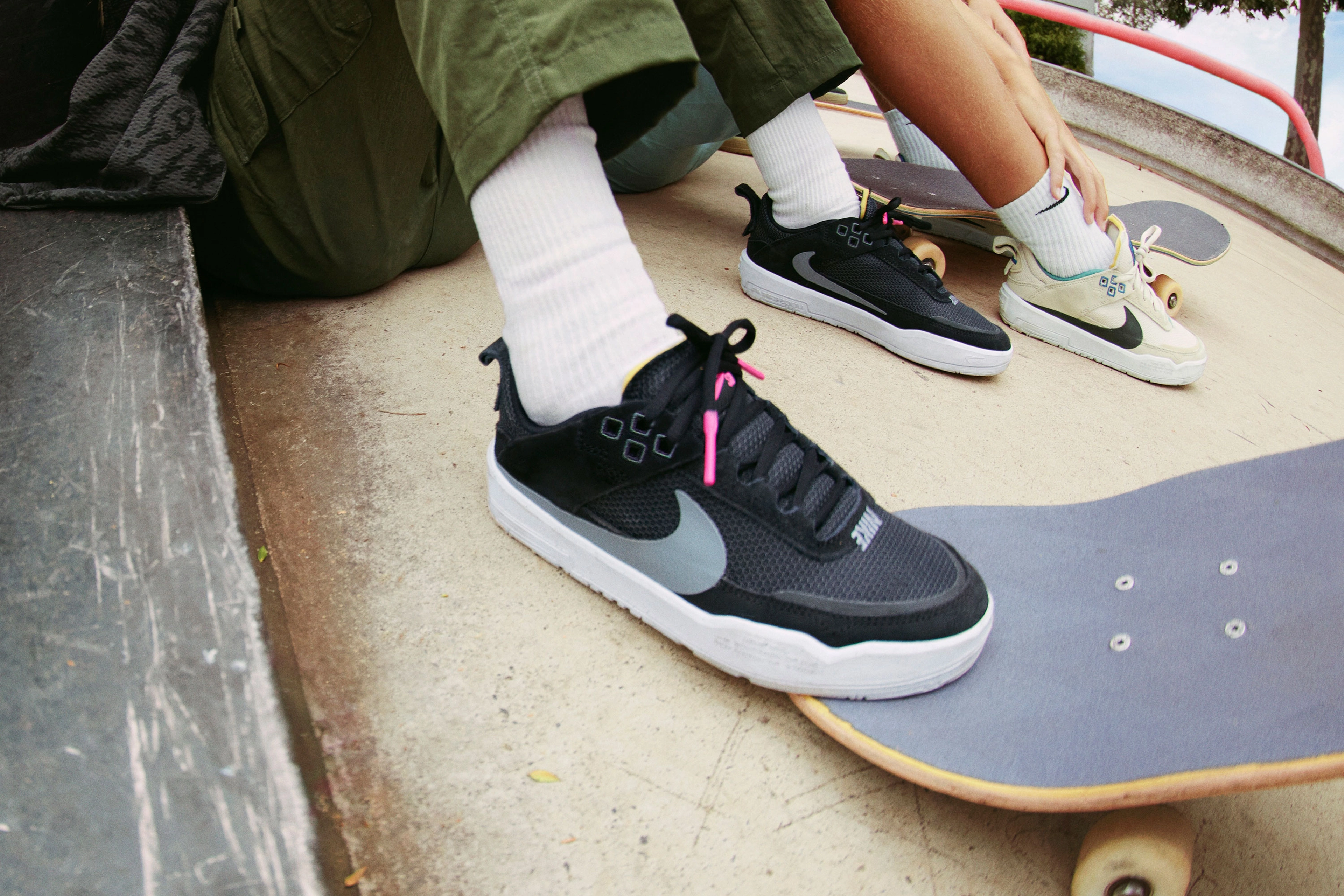 Close up view of two kids legs sitting down at a skatepark and both wearing the Nike SB Day One. One pair is black and the other pair is sanddrift.