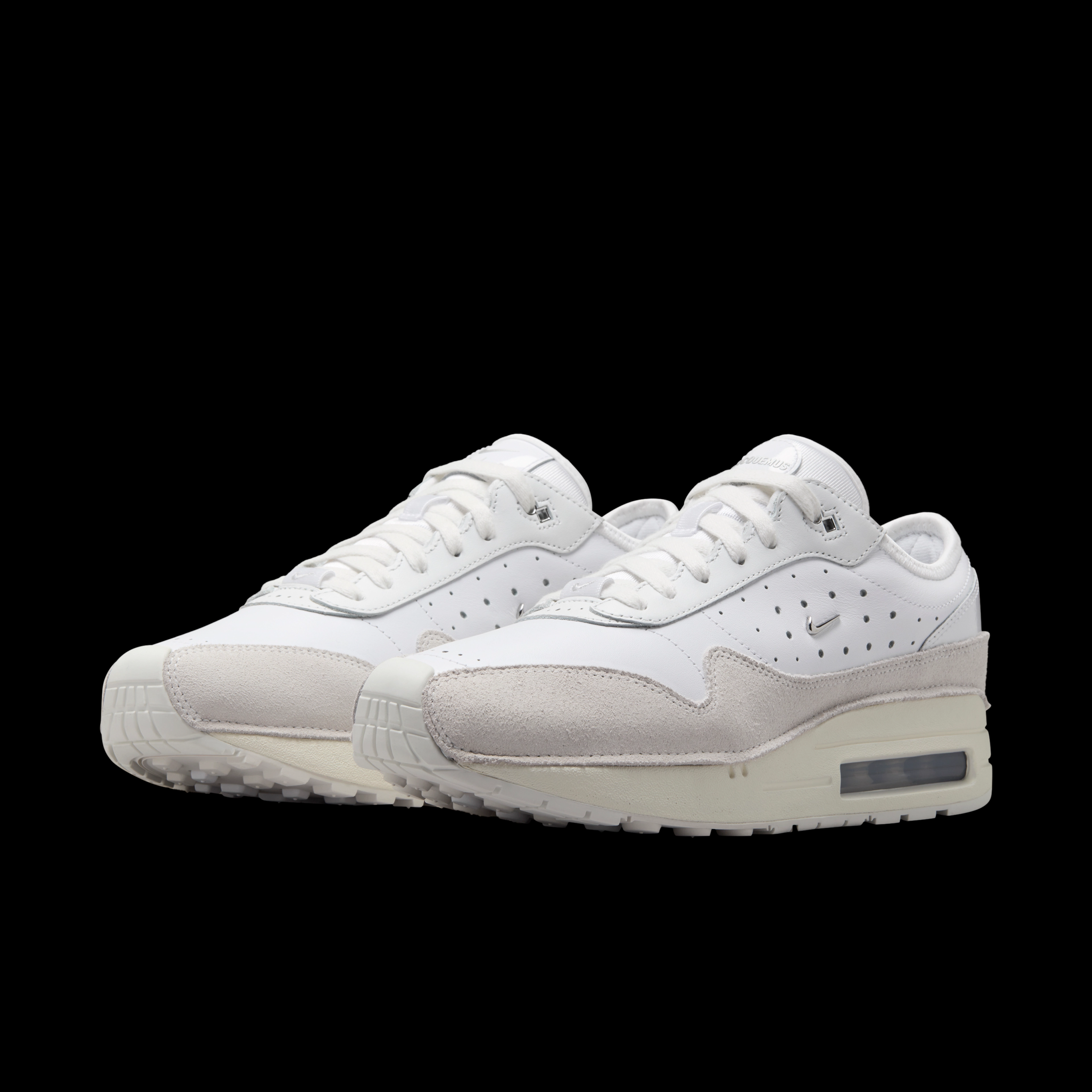 Front angled view of a pair of Nike x Jacquemus Air Max 1 sneakers. The shoes have a white upper with sail suede details, a white midsole, and white sole. There is a small metallic silver Swoosh on the side of the shoe. The pair floats on a black background.