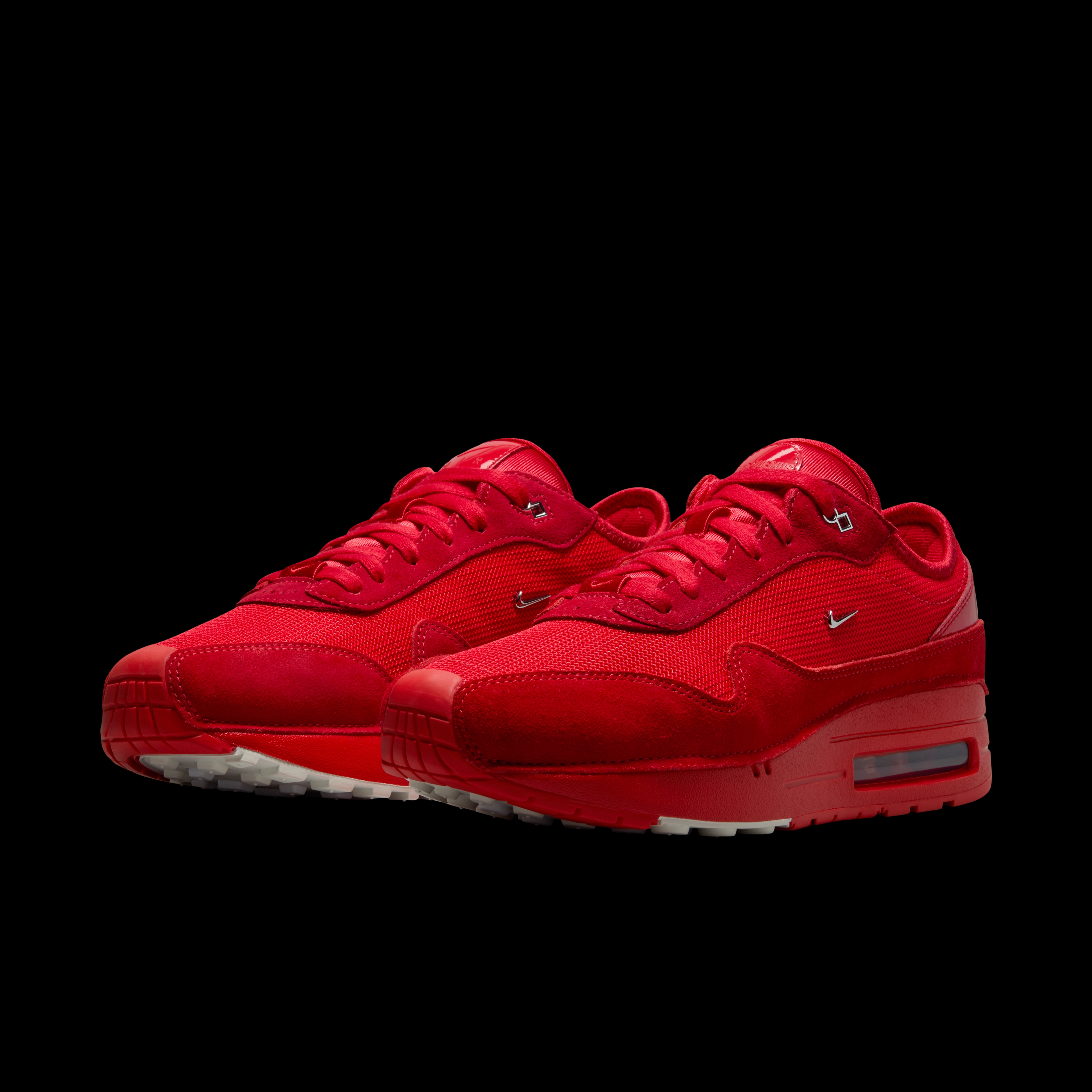 Front angled view of a pair of Nike x Jacquemus Air Max 1 sneakers in Mystic Red. The shoes have a red upper with red suede details, a red midsole, and a white and red sole. There is a small metallic silver Swoosh on the side of the shoe. The pair floats on a black background.