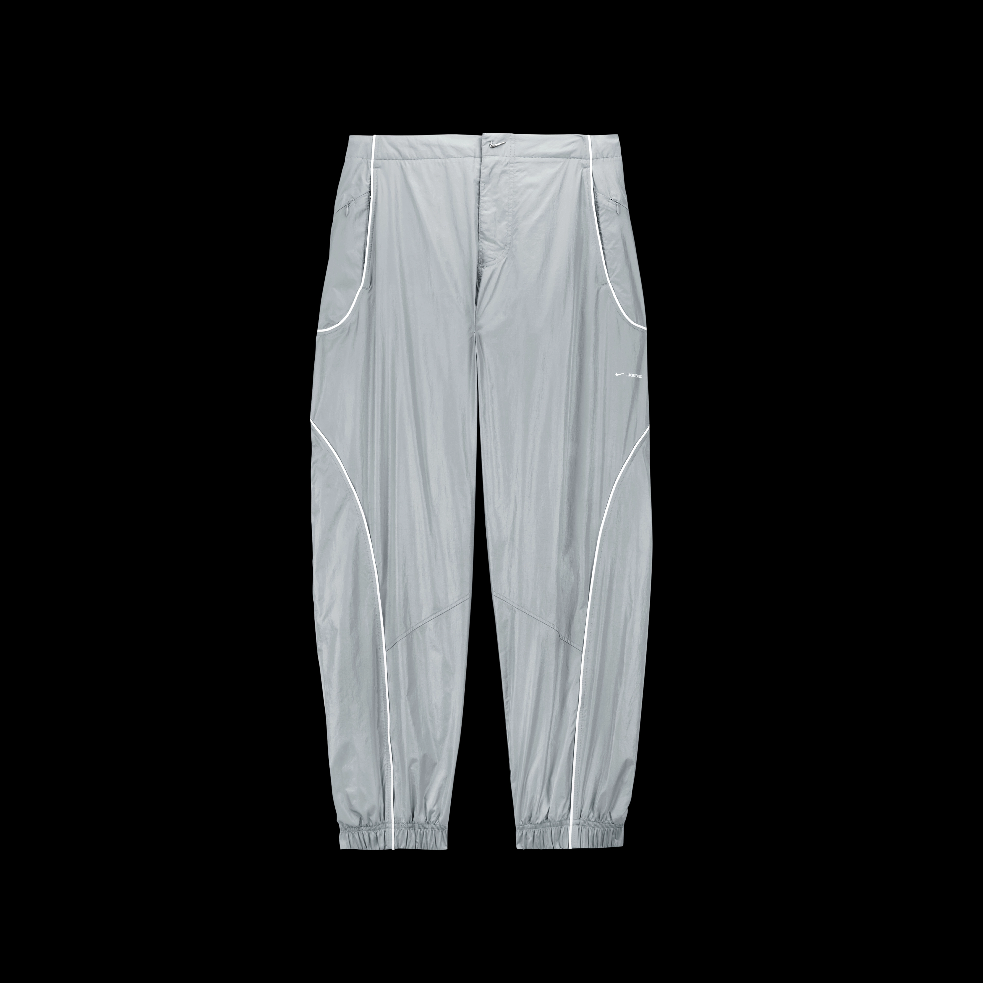 Overhead view of the Nike x Jacquemus Track Pant in particle grey with curved white pinstripes at the pocket and down the leg.  There is a small shiny silver swoosh as a closure on the waist, and elastic cuffs at the ankle. The pant floats on a black background.