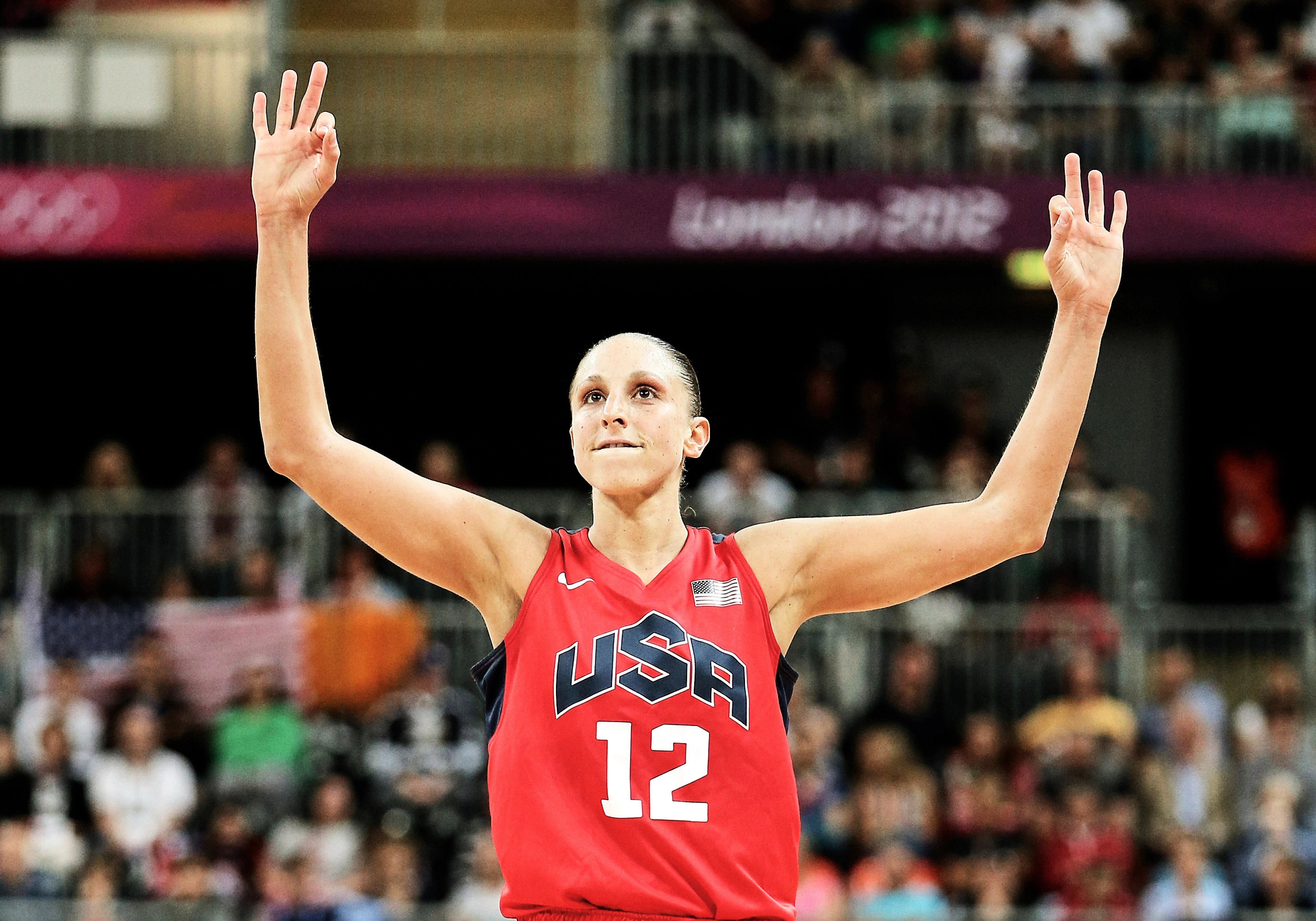 Diana Taurasi holds her hands high, with six fingers raised, signaling her 6 gold medals. Diana wears a read Team USA jersey, and a crowd looks on behind her.