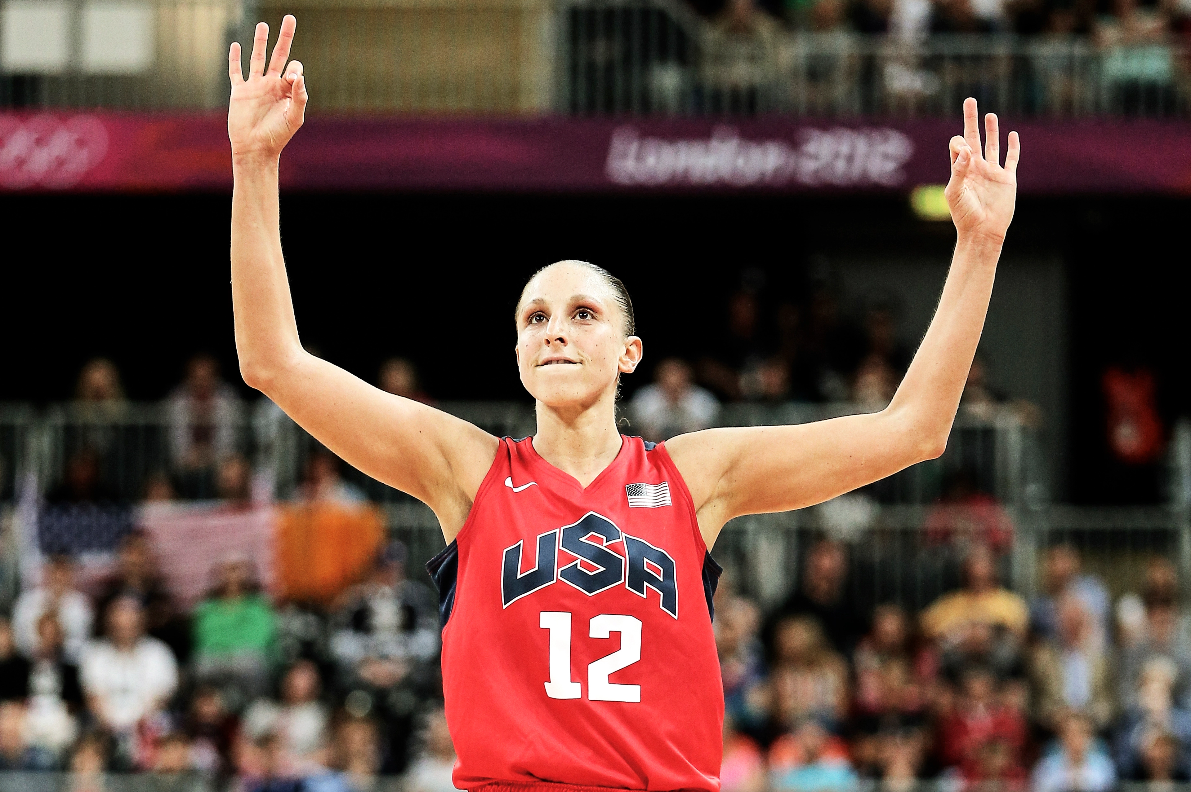 Diana Taurasi holds her hands high, with six fingers raised, signaling her 6 gold medals. Diana wears a read Team USA jersey, and a crowd looks on behind her.