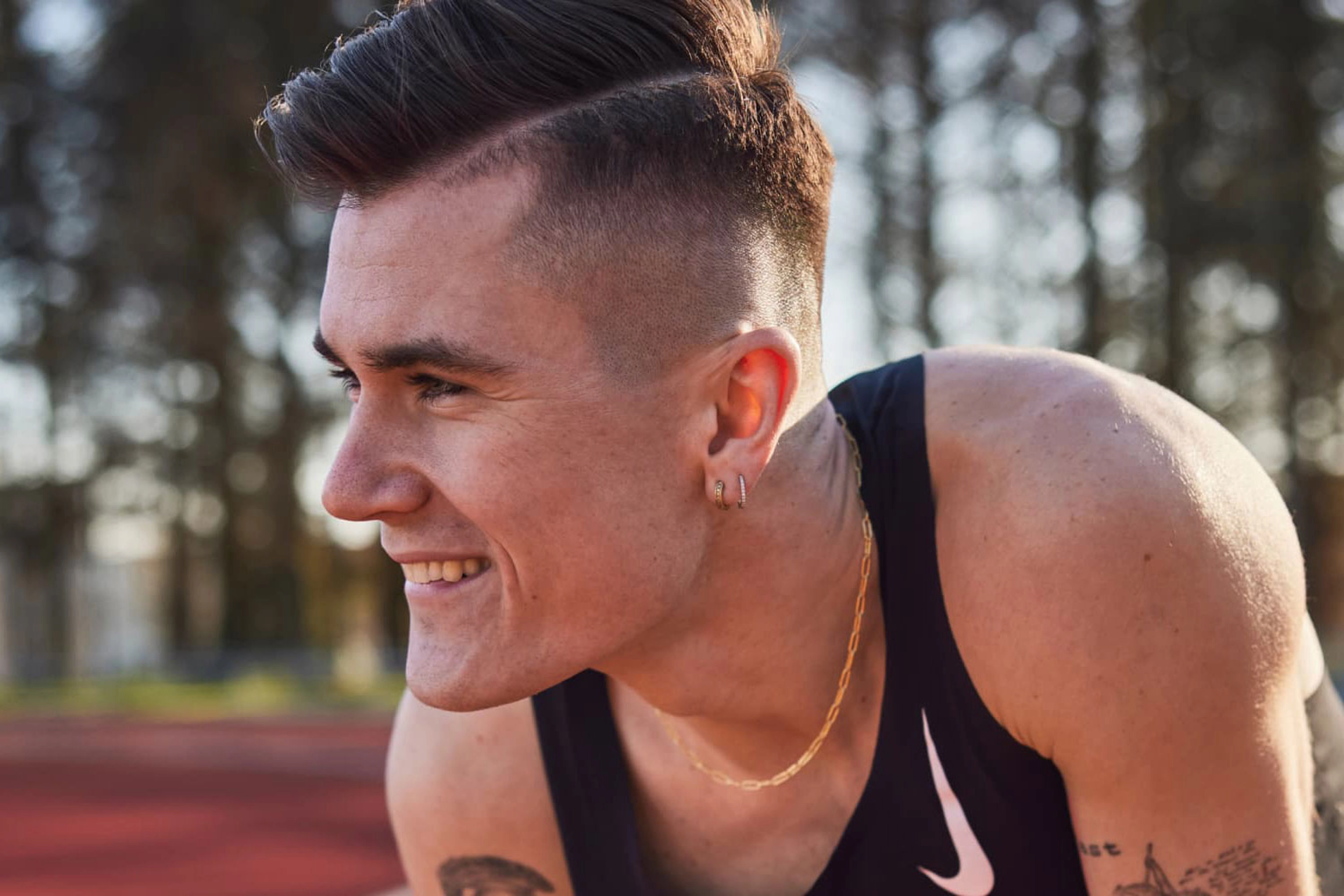 Jakob Ingebrigtsen, crouches on a track, and smiles as he looks to the left, wearing a black Nike singlet and a gold chain around his neck.