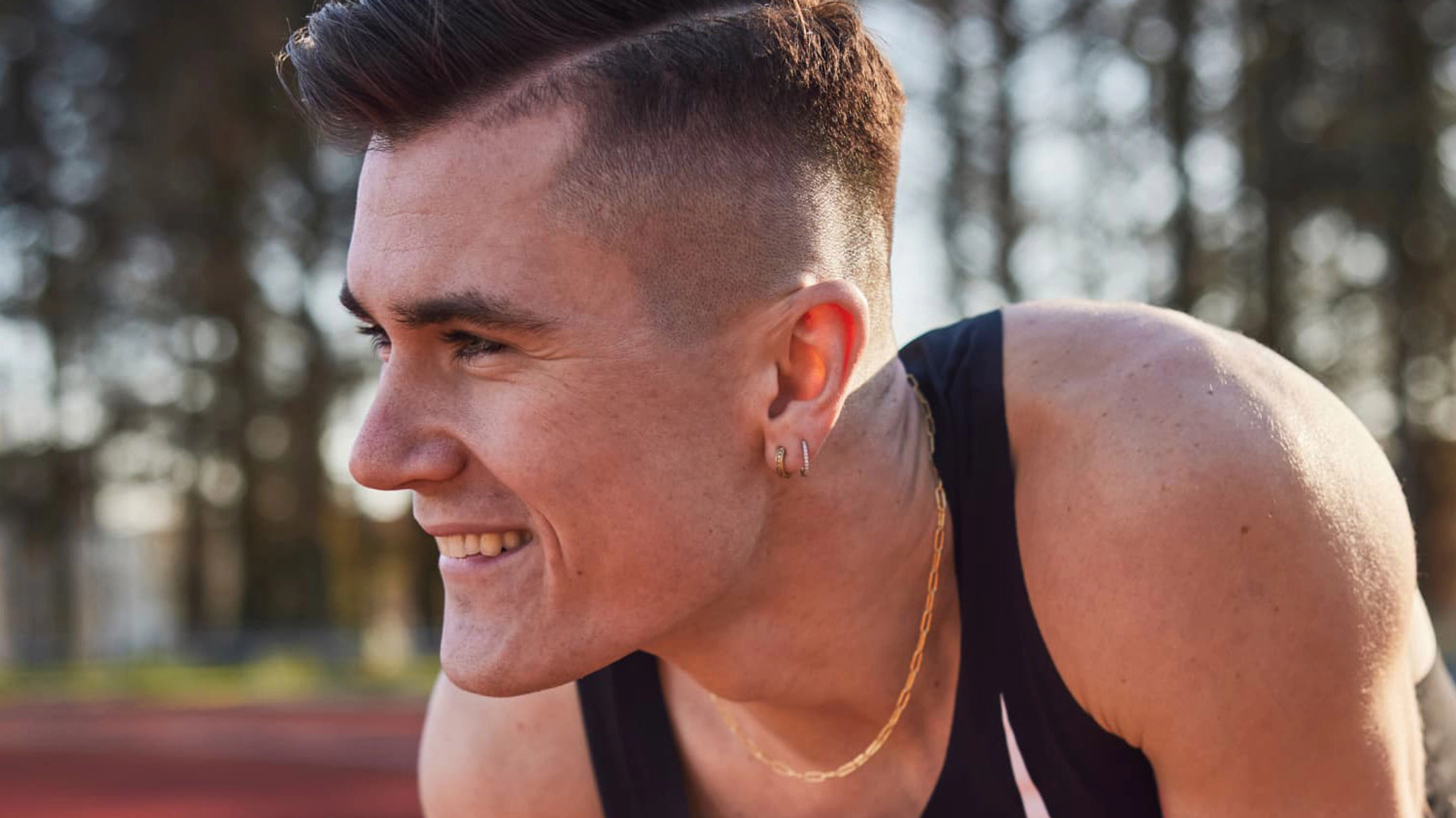 Jakob Ingebrigtsen, crouches on a track, and smiles as he looks to the left, wearing a black Nike singlet and a gold chain around his neck.