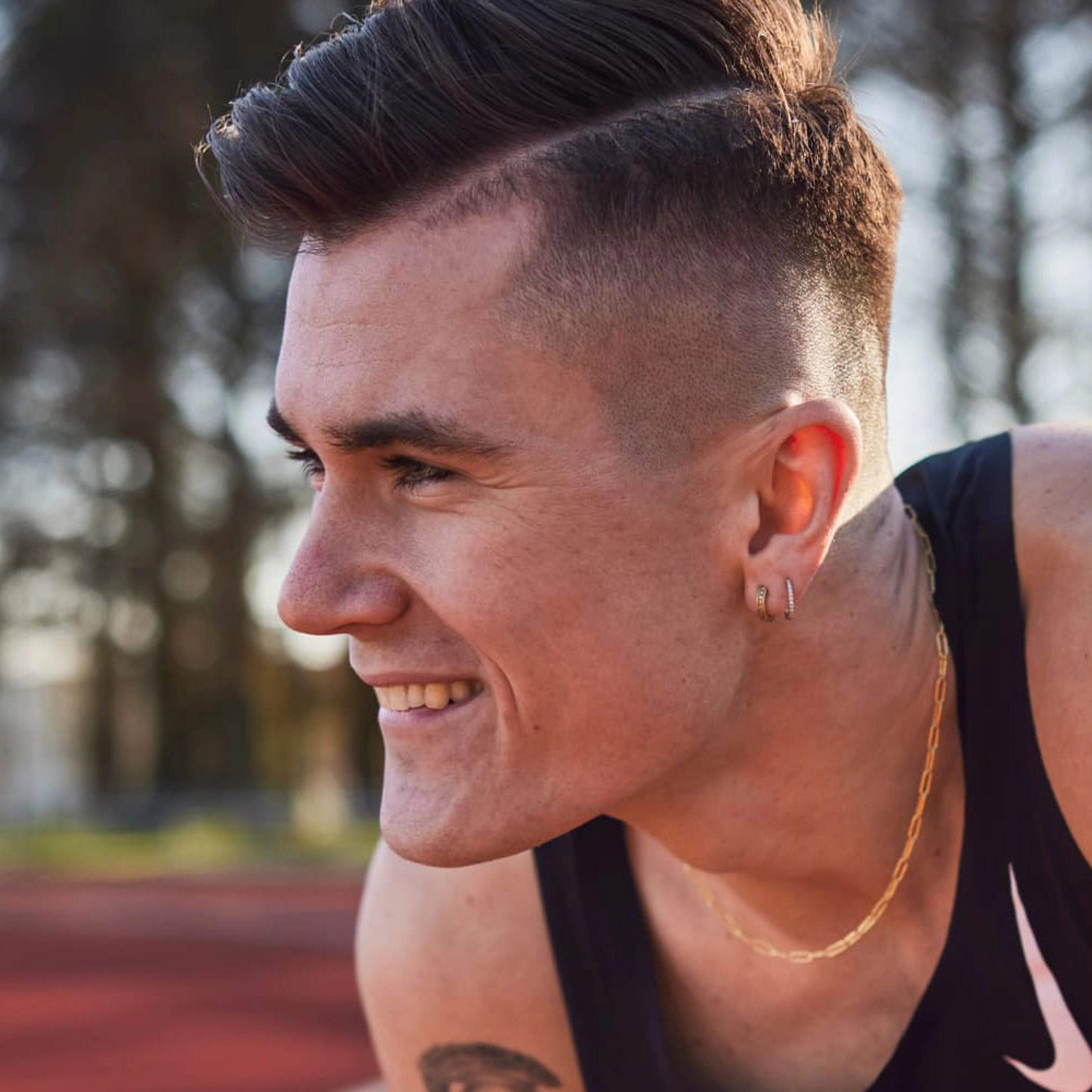 Jakob Ingebrigtsen, crouches on a track, and smiles as he looks to the left, wearing a black Nike singlet and a gold chain around his neck.