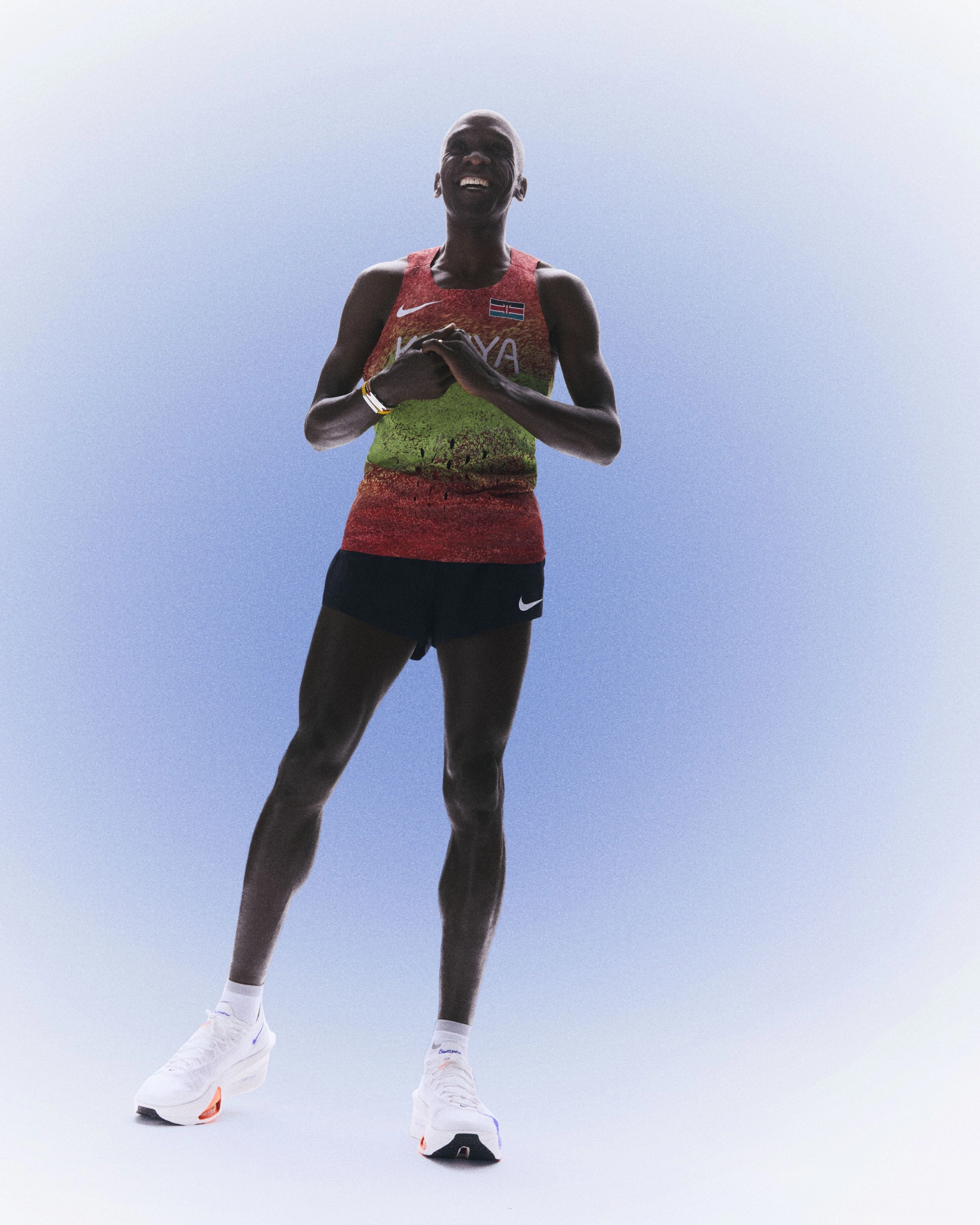 Front full body view of Eliud Kipchoge smiling while wearing his Kenya track and field kit uniform.