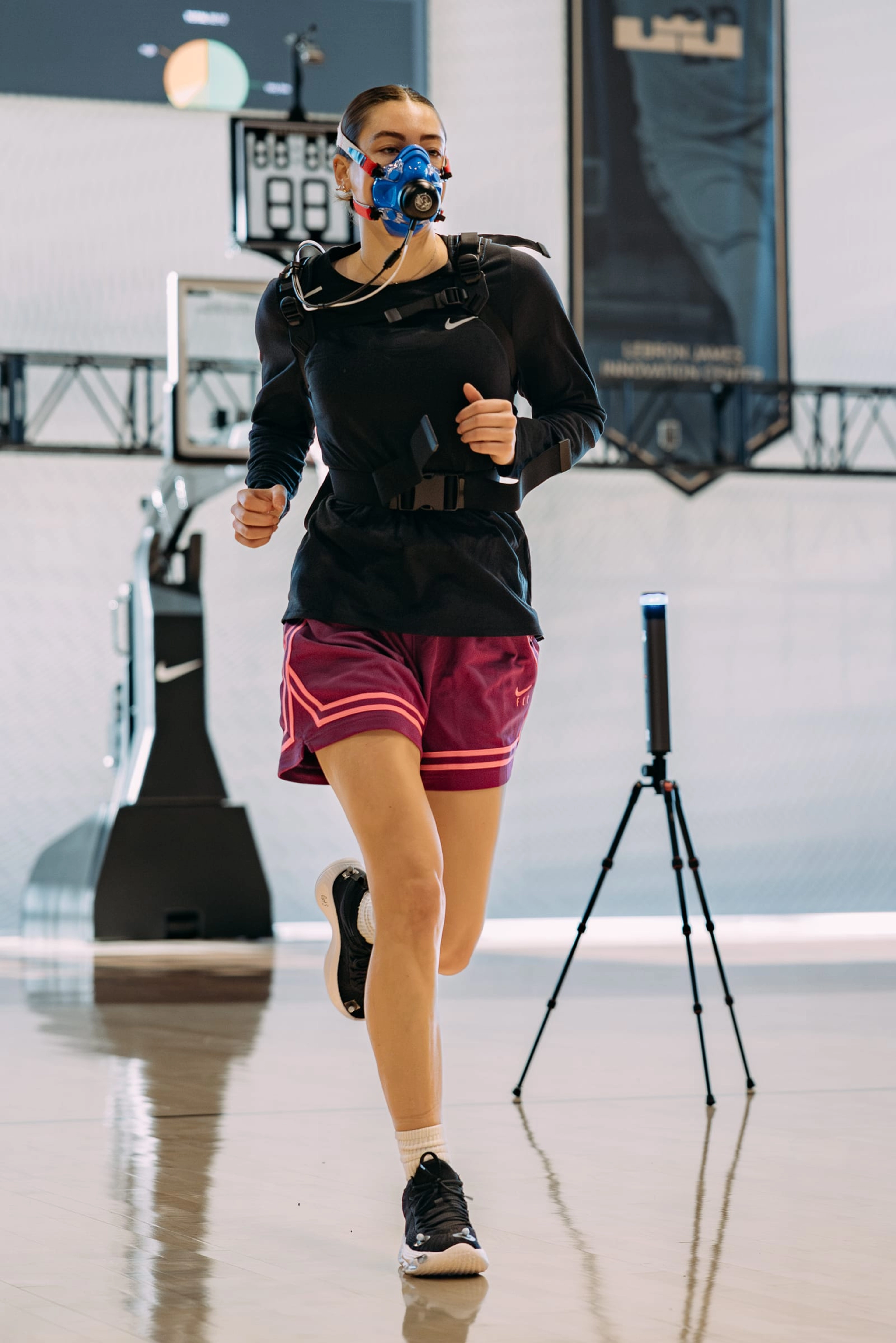 A female basketballer, wearing burgundy shorts and a long-sleeve black top, runs down the basketball court in the Nike GT Hustle 3, during testing in the Nike Sports Research Lab. She wears an oxygen mask and sensors, and there is scientific equipment in the background.