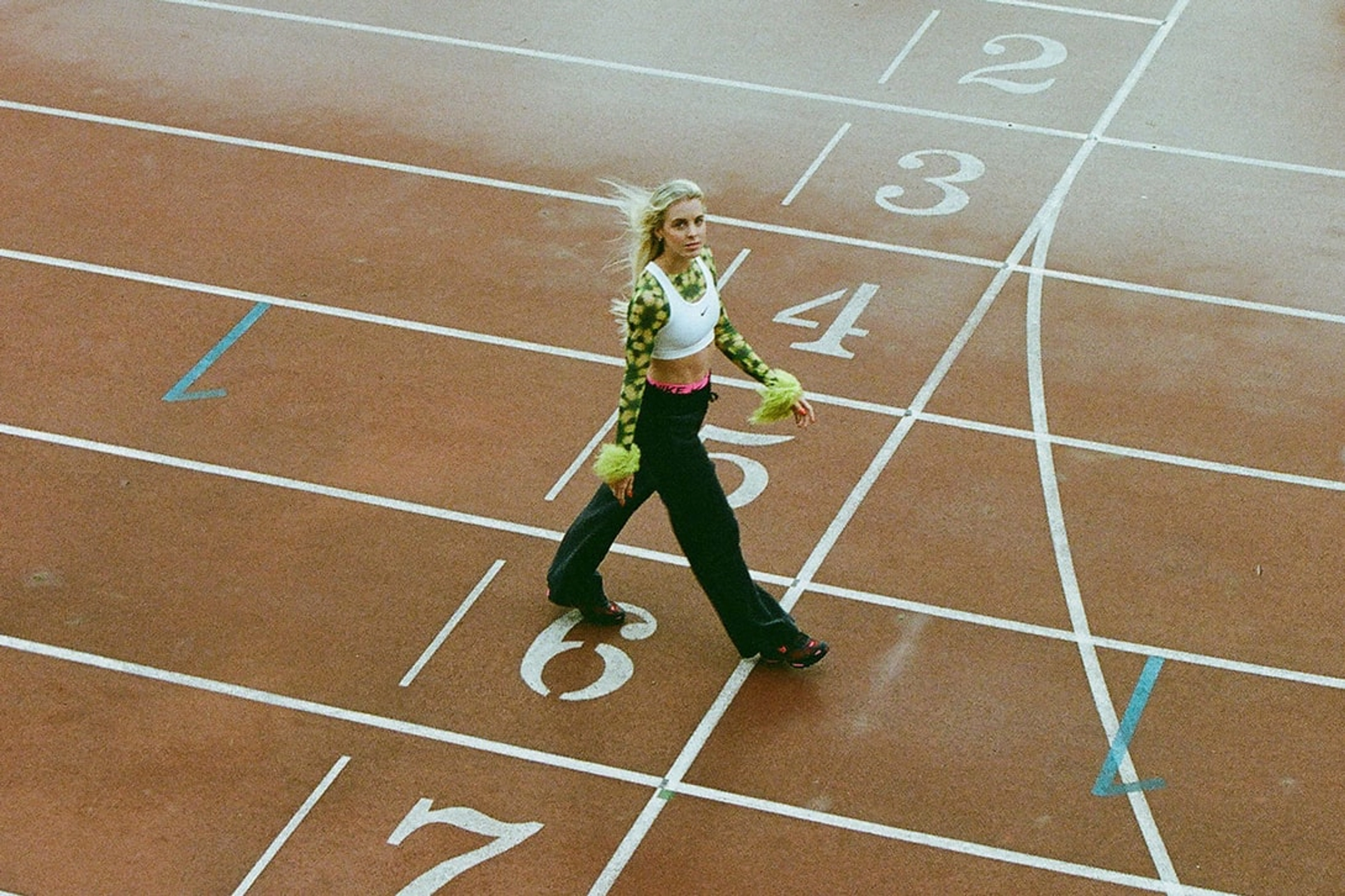Overhead view of Keely Hodgkinson walking down the #6 Track lane wearing a white Nike Swoosh bra over a green and black crop top and black pants with the pink waistband of Nike Pro shorts showing. She looks up towards the camera, with her hair blowing in the wind behind her.
