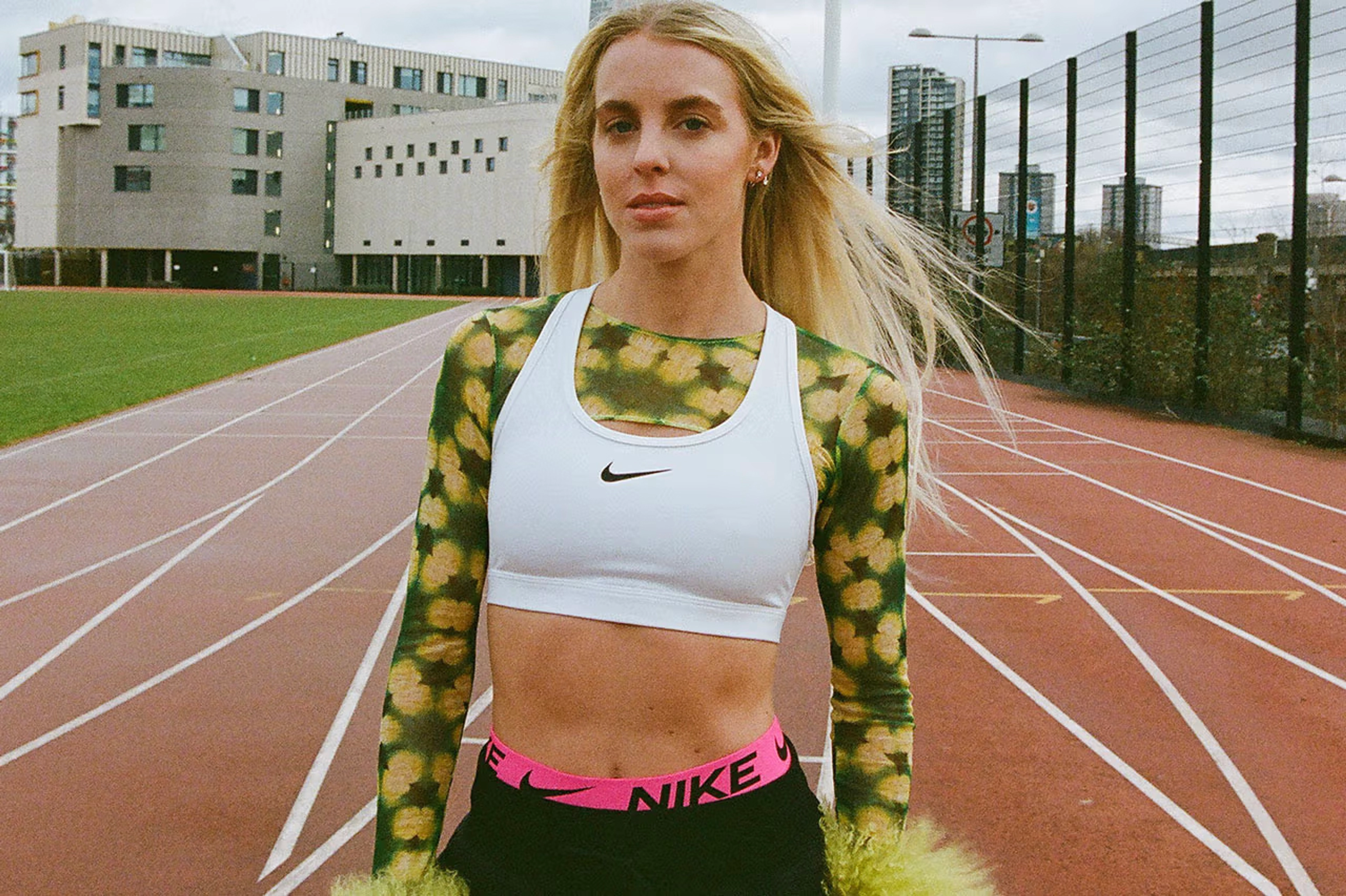 Close up of Keely Hodgkinson standing on a Track. She wears a white Swoosh bra layered over a green and black crop top, and black pants with the pink band of Nike Pro shorts visible. A cityscape is visible in the background.