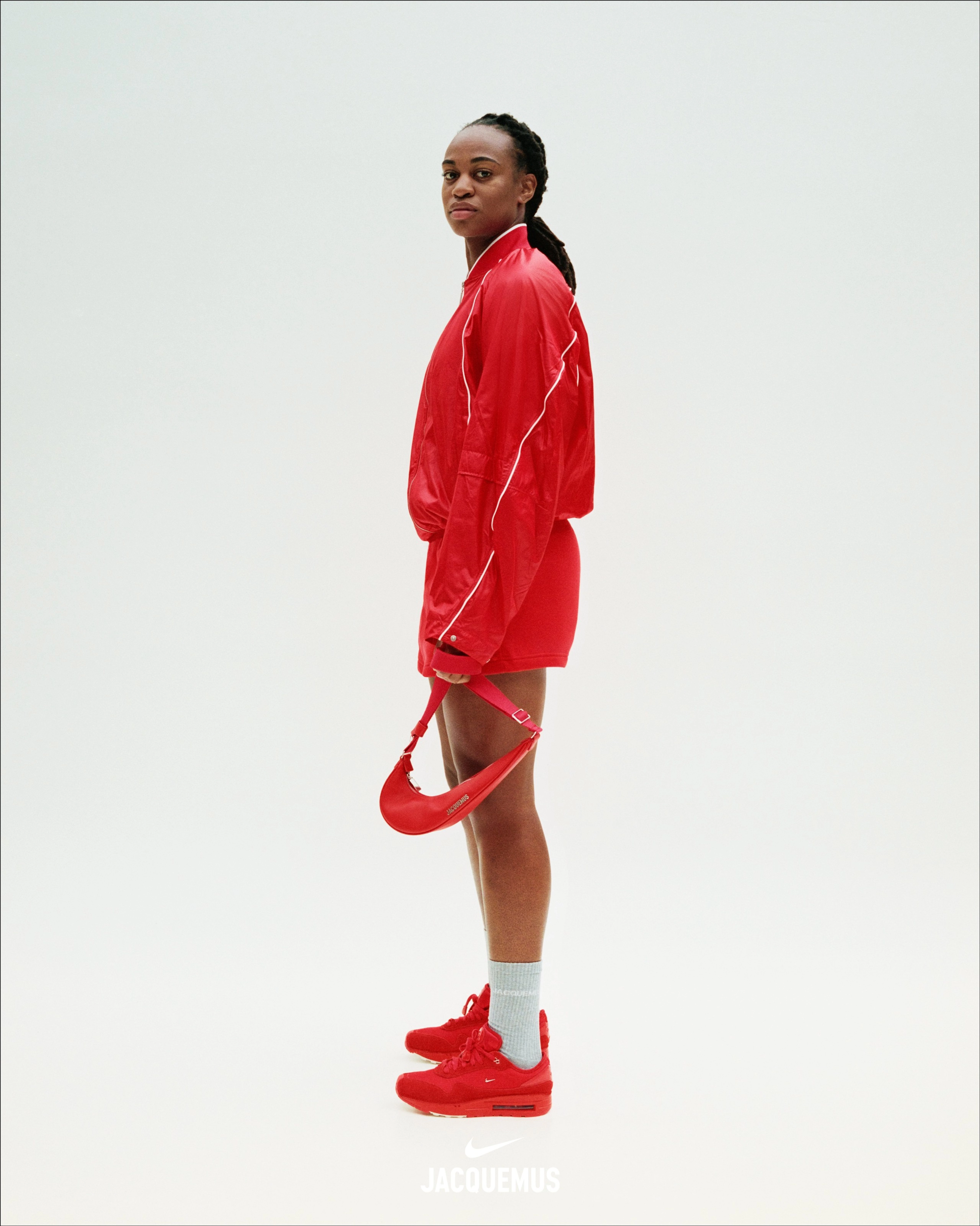 Side view of Marie-Antoinette Katoto wearing a red Nike x Jacquemus Track Jacket, with red shorts, light grey socks, and the Nike x Jacquemus Women's Air Max 1 '86 Big Bubble in Mystic Red. Marie-antoinette faces left as she stands on a white background, looking at the camera and holding a red Nike x Jacquemus Le Swoosh Crossbody Bag in her hand.