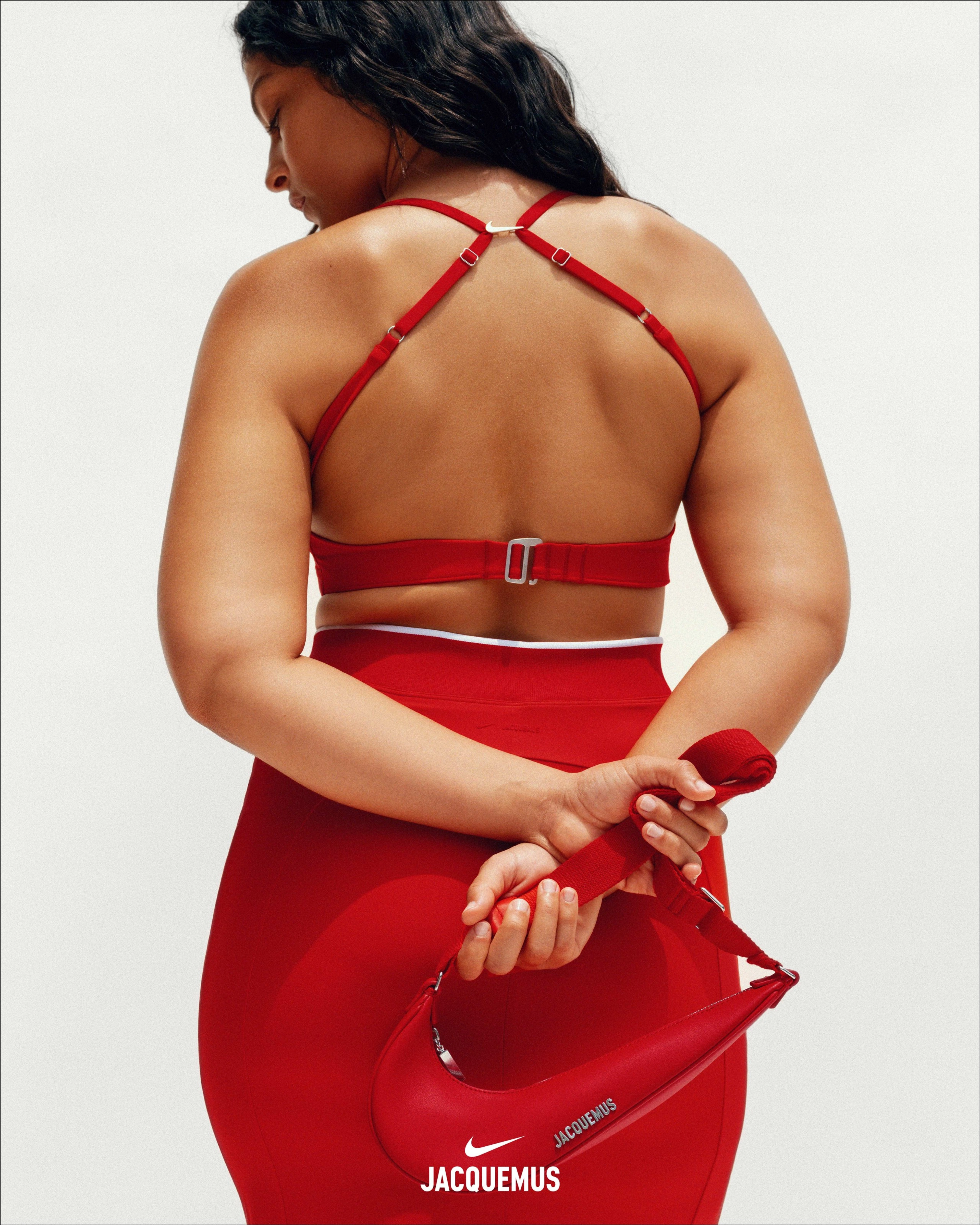 Back view of a female athlete, who is holding her arms behind her back as she holds a Nike x Jacquemus Le Swoosh Crossbody Bag in red, in one hand. We see the back of the Nike X Jacquemus Wome's Skirt and Women's Bra is red. The woman looks to the left, as she stands against a white background.