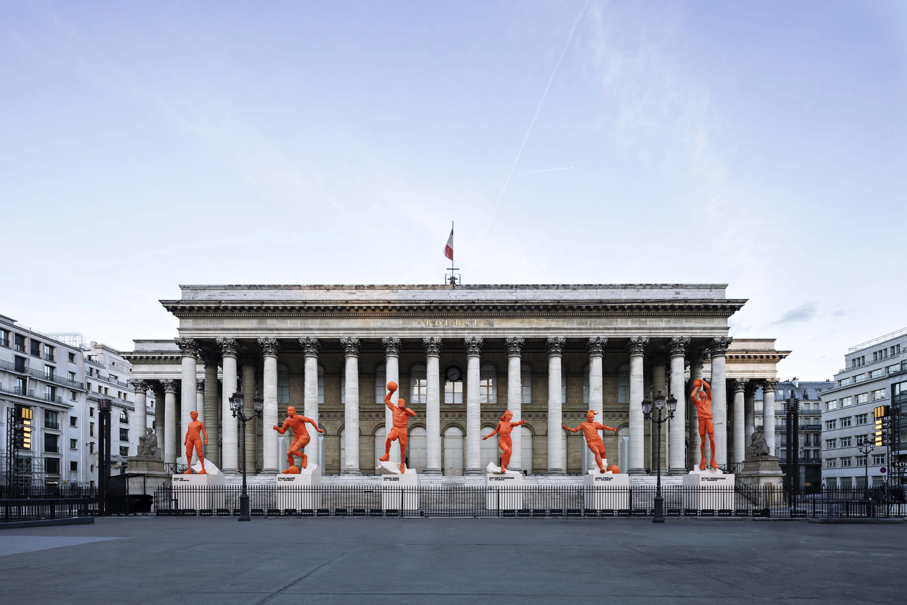 Street view photo of the Palais Brongniart building with 6 orange statues in athletic poses in front of it.