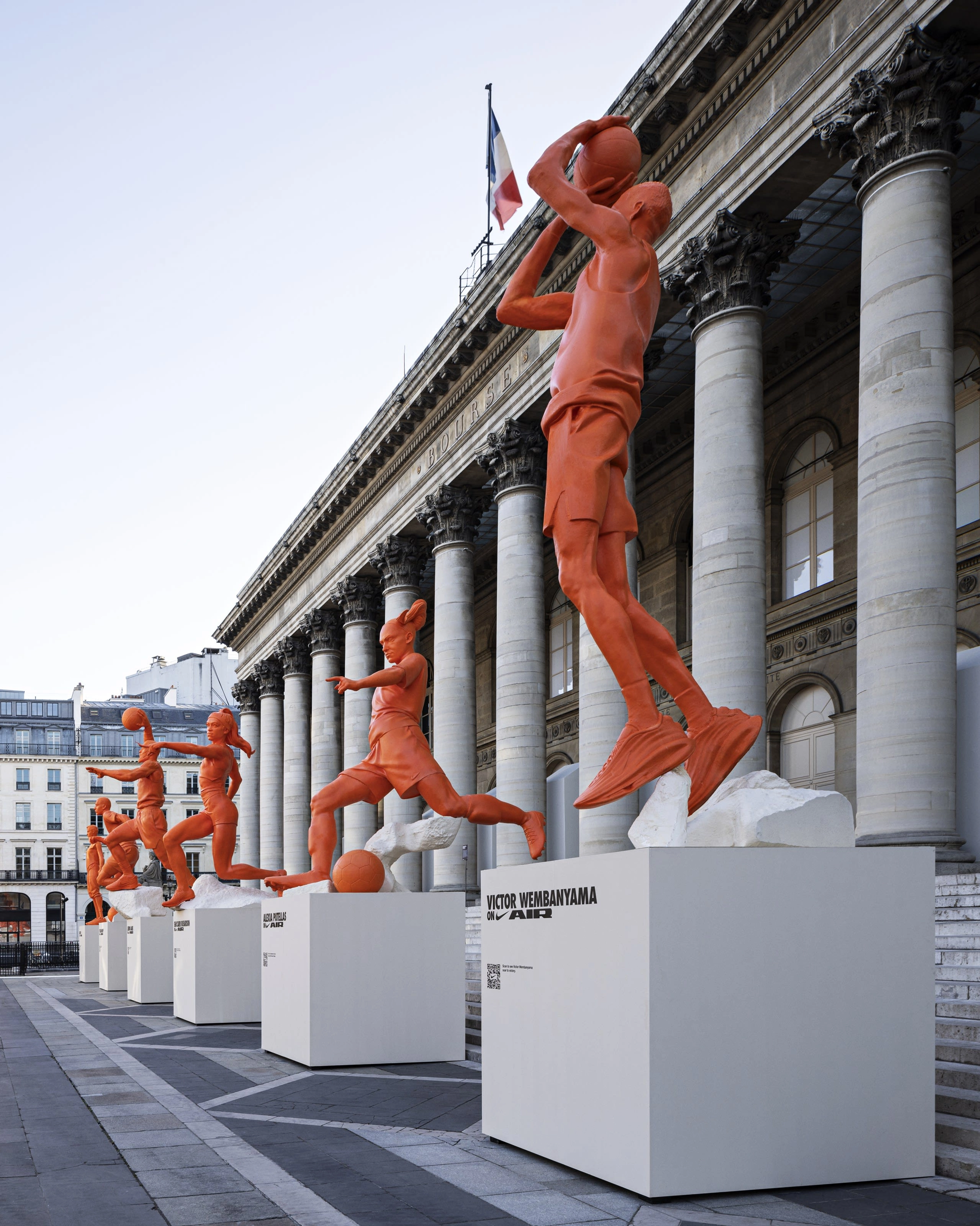 Side angle view of the 6 orange athlete statues in front of the Palais Brongniart building. The nearest statue is observably a replica of Victor Wembanyama shooting a basketball.