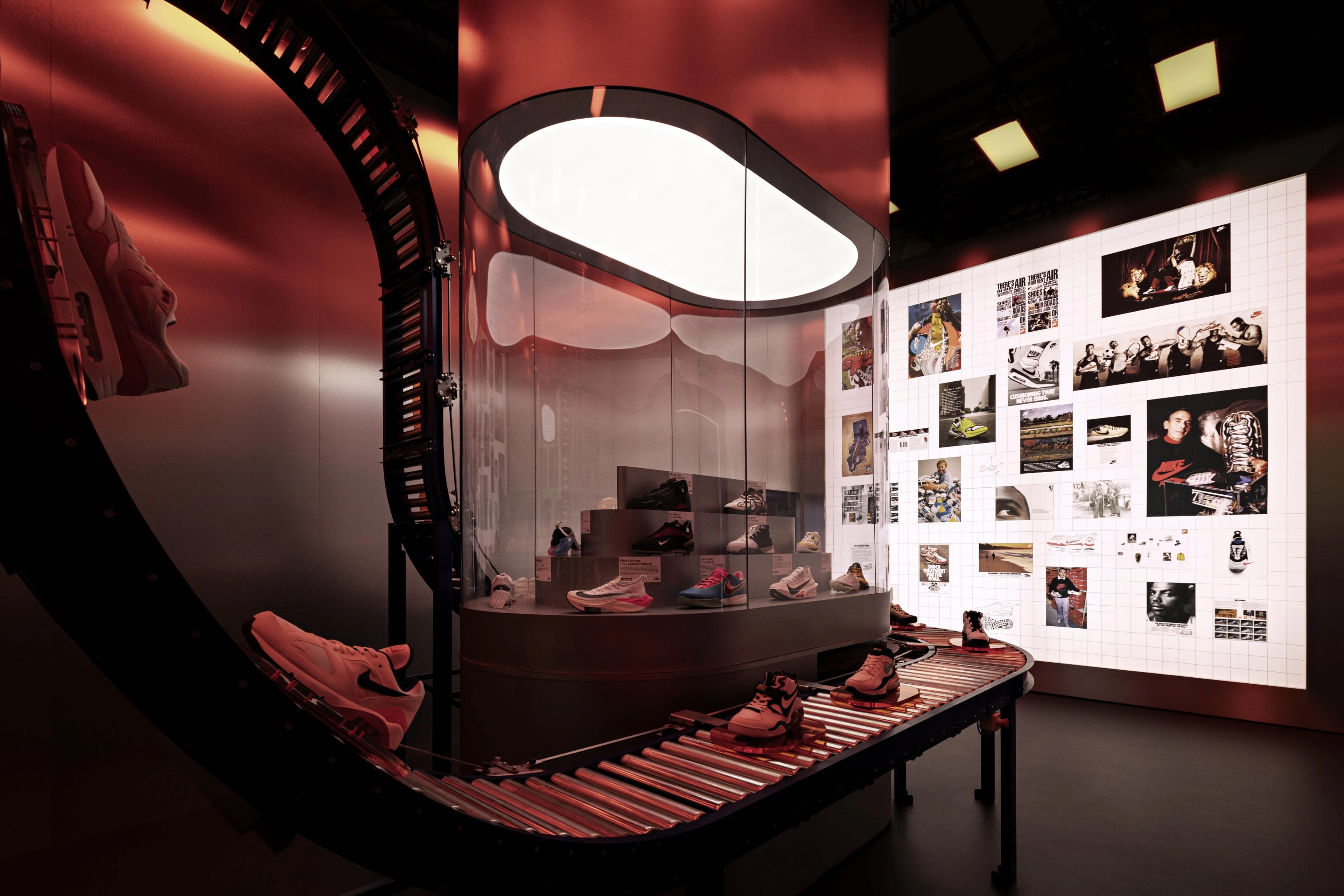 A dimly lit room featuring an artistic display of Nike shoes on a conveyor belt in the center and a backlit photo wall behind it.