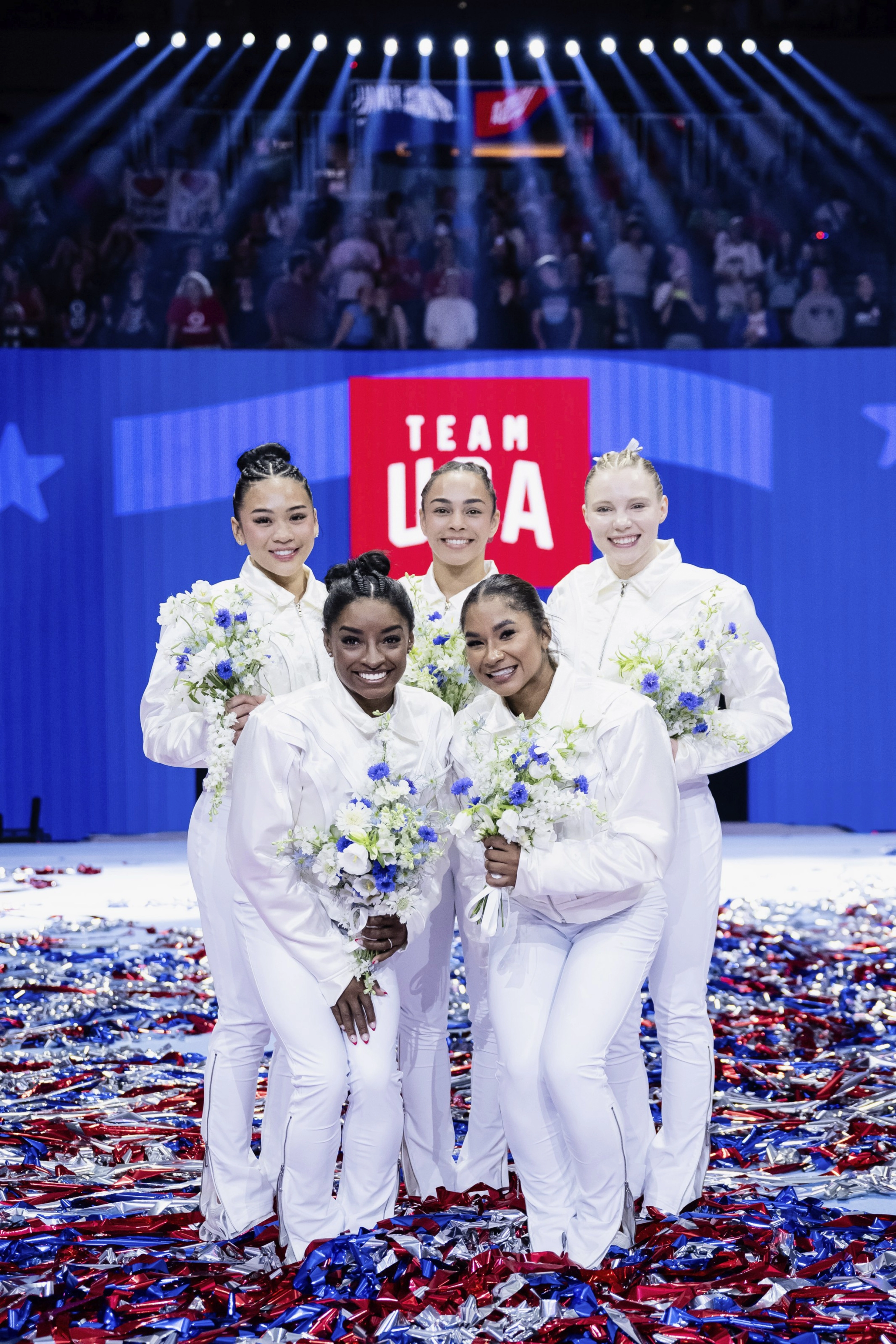 Jordan Chiles stands with Team USA Women’s Gymnastics. The team wears matching white outfits and holds bouquets of white and blue flowers. A red and white Team USA signs sits behind them, and the floor is covered with shiny red, white and blue streamers.