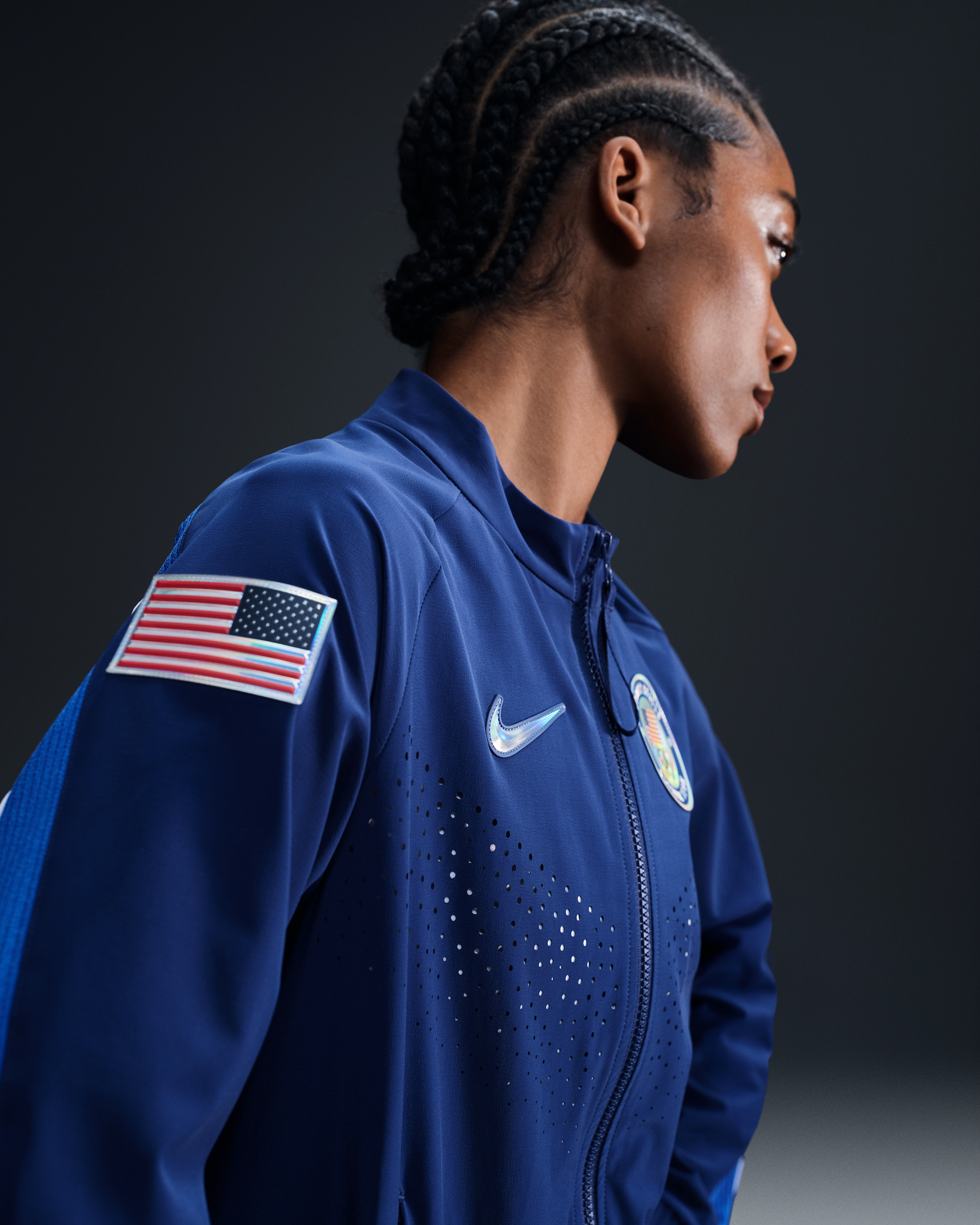 Side view of a female athlete wearing the Women's Medal Ceremony Jacket in the exclusive Team USA bright blue colorway. A USA flag patch is sewn into the upper arm, and the jacket has a silver iridescent Swoosh. The athlete stands against a dark grey background.