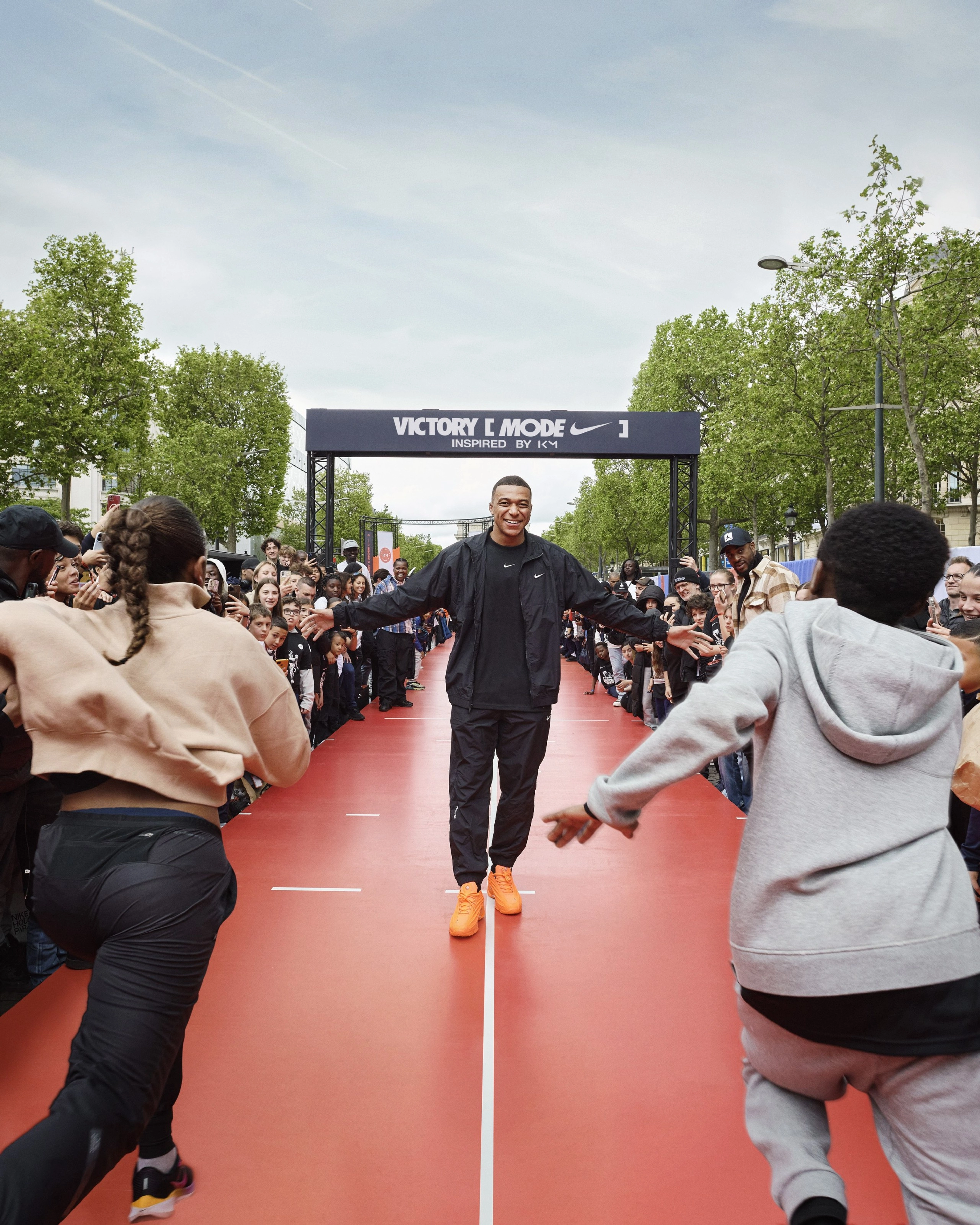 Front view of Kylian Mbappe, walking down the center of a stage that looks like track lanes. Runners in Nike product give him high fives as they pass. Mbappe is in black Nike tee, jacket and pants, and is smiling at the runners as they pass.