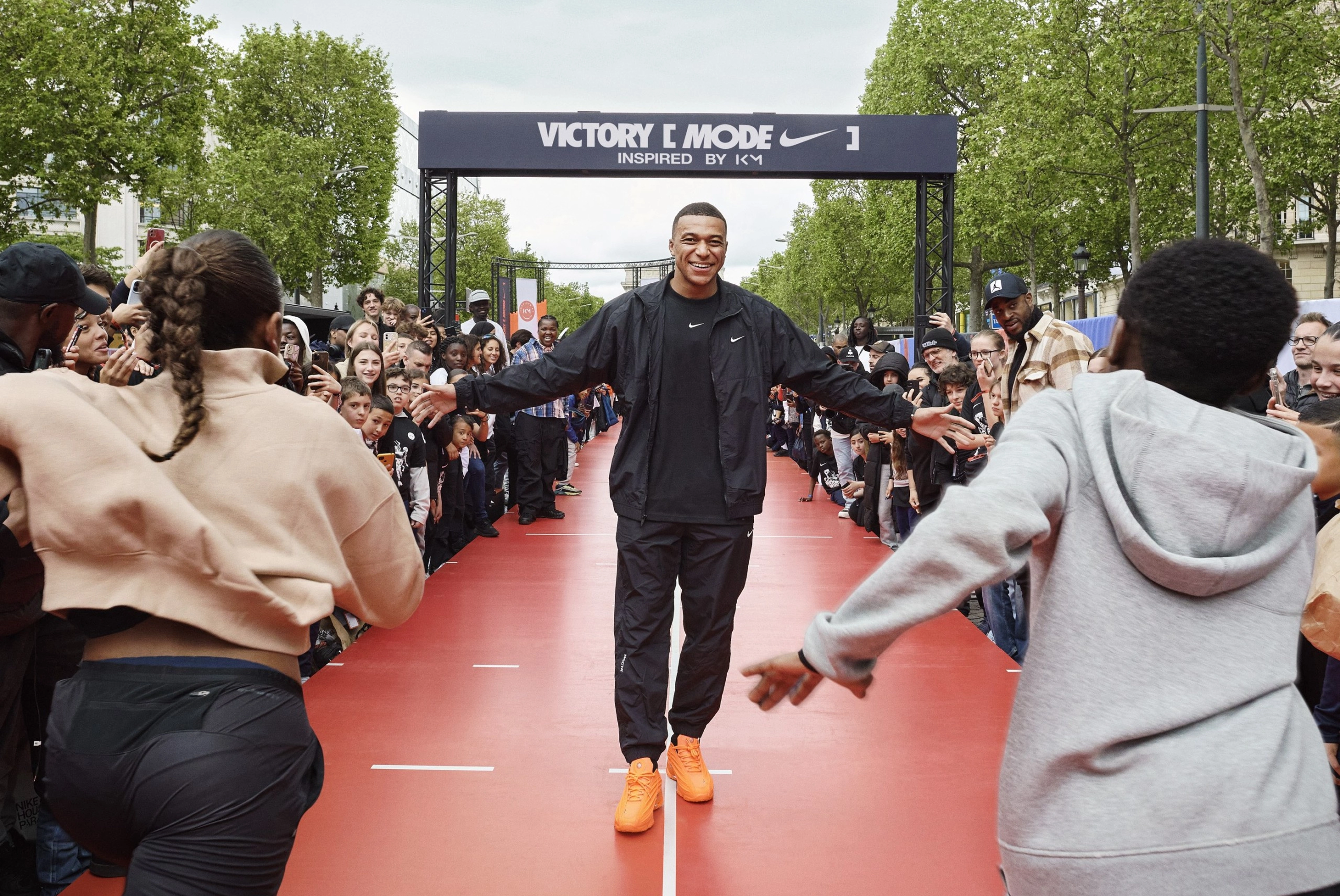 Front view of Kylian Mbappe, walking down the center of a stage that looks like track lanes. Runners in Nike product give him high fives as they pass. Mbappe is in black Nike tee, jacket and pants, and is smiling at the runners as they pass.