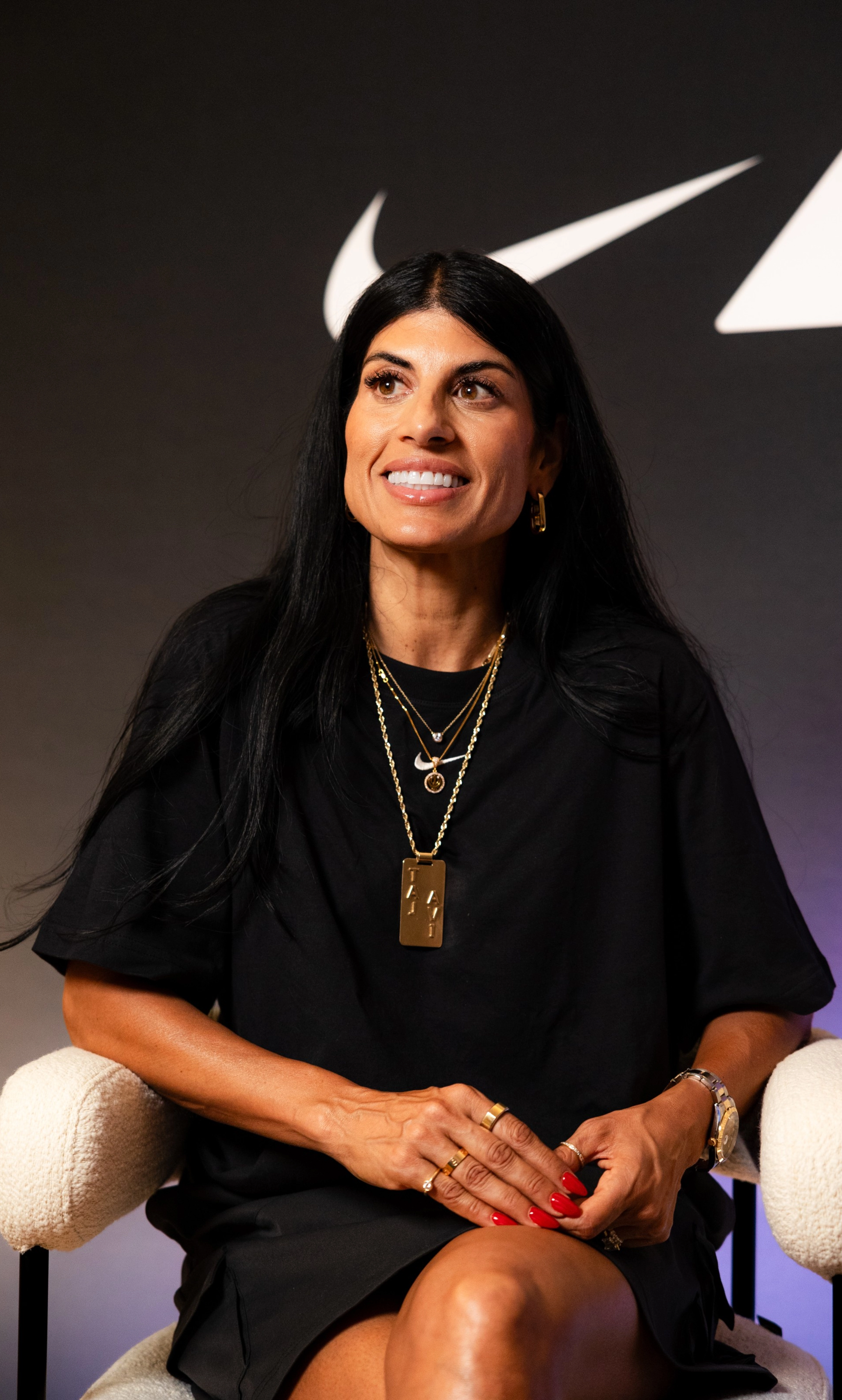 Close up of Nike track and field coach, Diljeet Taylor, wearing a black Nike Sportswear Chill Knit dress, with layered gold jewelry. She sits in a white chair, against a dark background, looking slightly to the left.