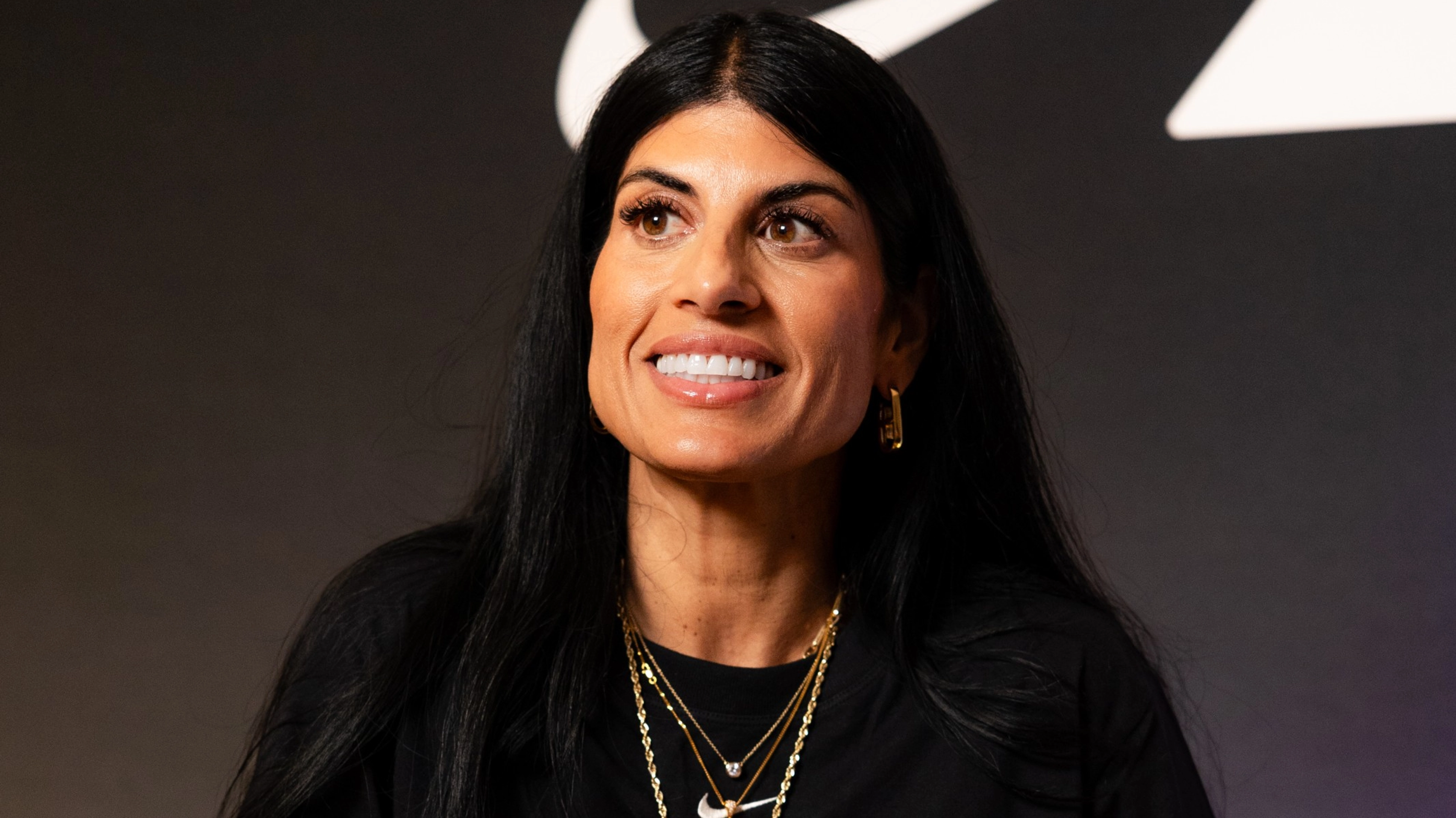 Close up of Nike track and field coach, Diljeet Taylor, wearing a black Nike Sportswear Chill Knit dress, with layered gold jewelry. She sits in a white chair, against a dark background, looking slightly to the left.