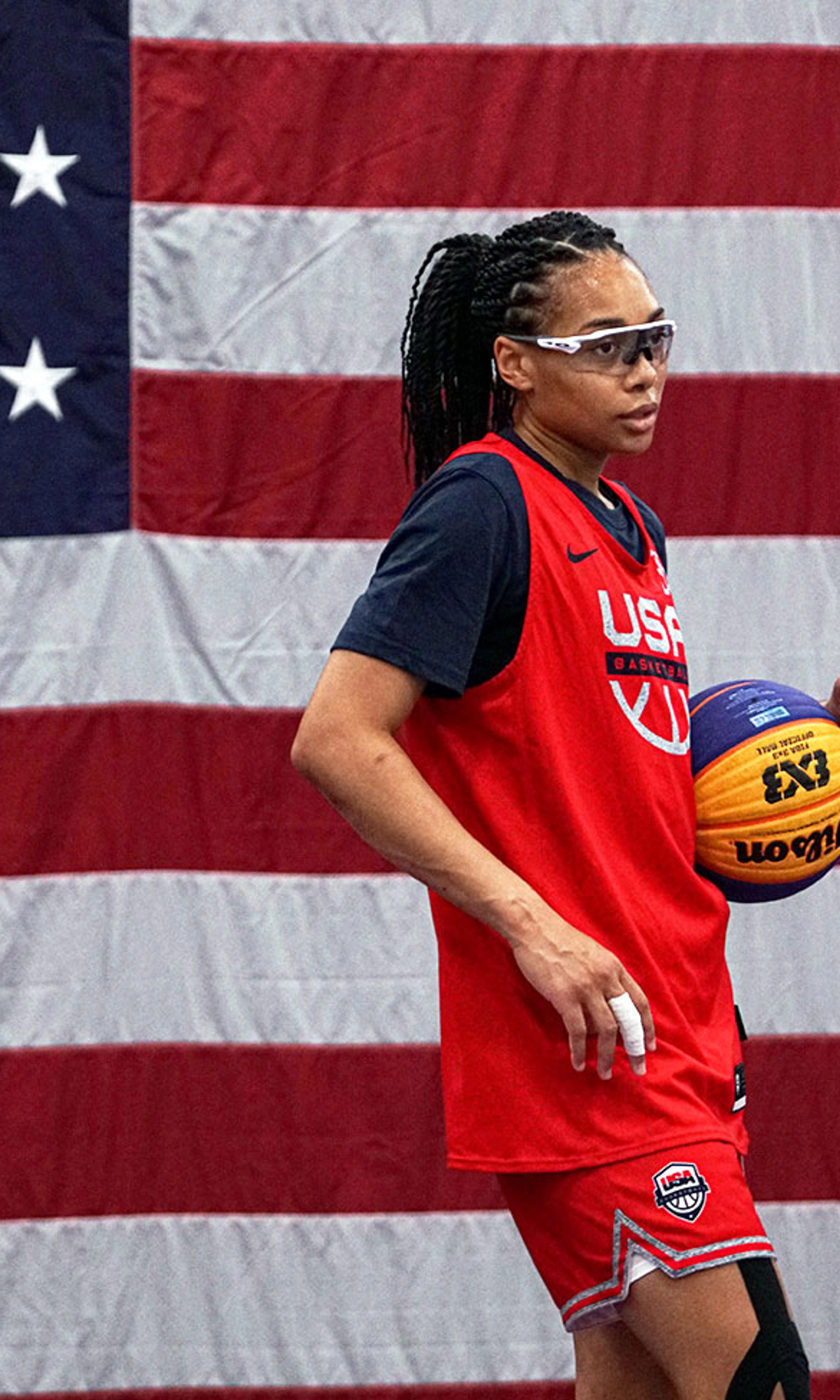 Allisha Gray wearing safety glasses and a red Nike USA basketball jersey, holding a basketball with a US flag in the background.