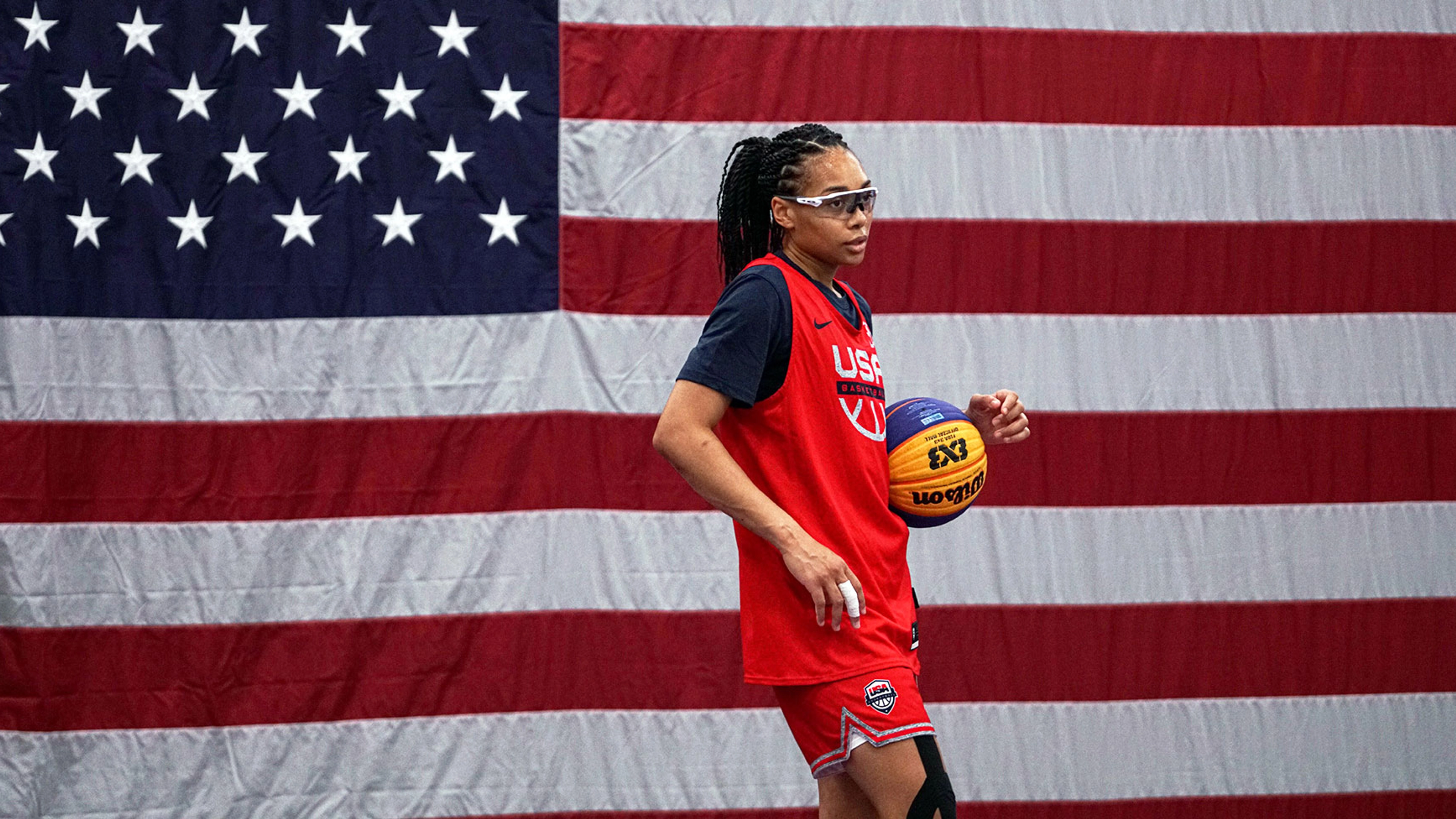 Allisha Gray wearing safety glasses and a red Nike USA basketball jersey, holding a basketball with a US flag in the background.