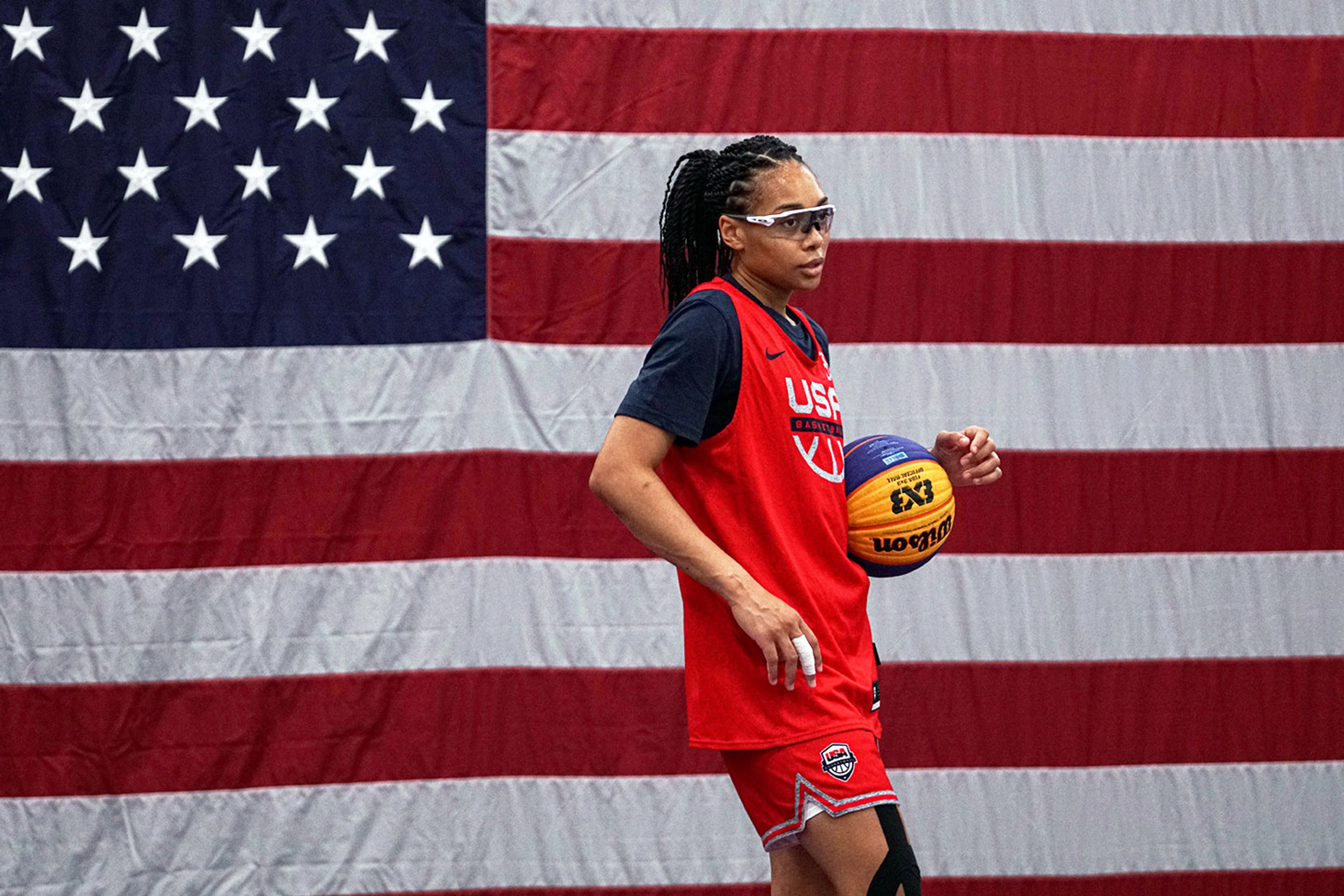 Allisha Gray wearing safety glasses and a red Nike USA basketball jersey, holding a basketball with a US flag in the background.
