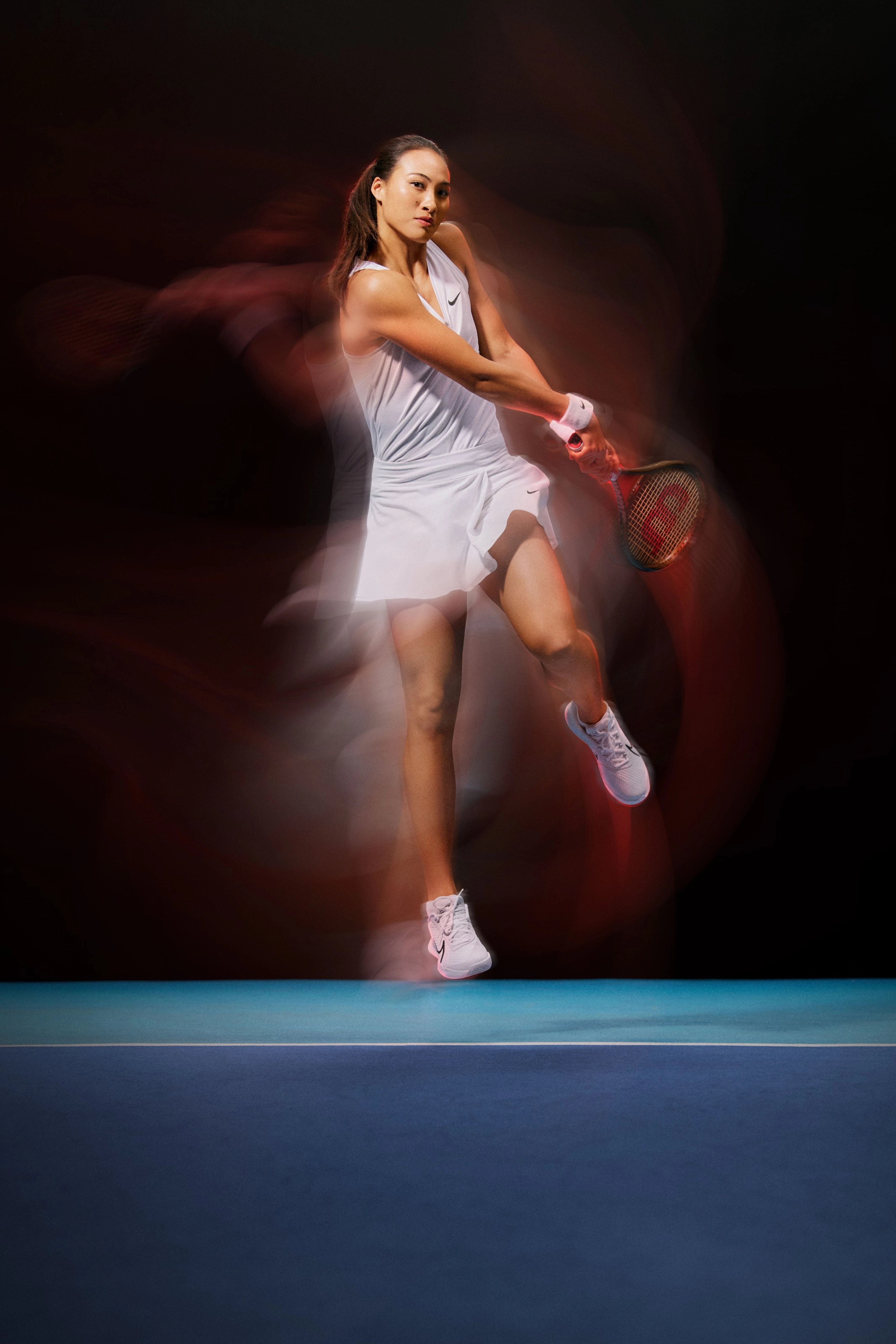 Full body view of Qinwen Zheng as she swings her tennis racquet, hovering in the air above a blue court. Qinwen wears the NikeCourt Victory tank in white, a white tennis skirt, and white court shoes with a black outlined Swoosh. There is a blur effect on the photo indicating the speed of her movement.